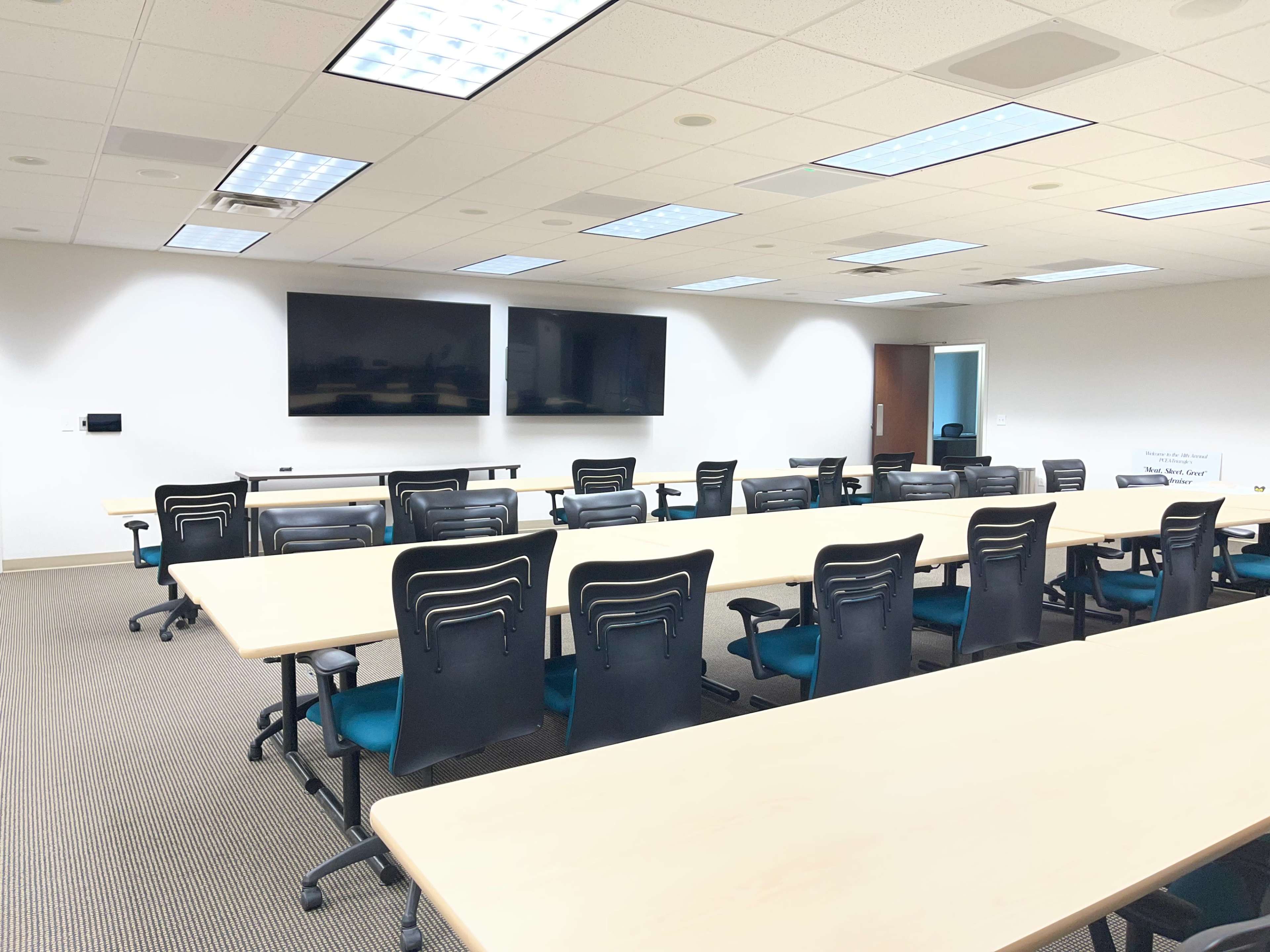 The image shows a modern conference room with multiple tables and chairs arranged for meetings, two wall-mounted screens, and a neutral color scheme.