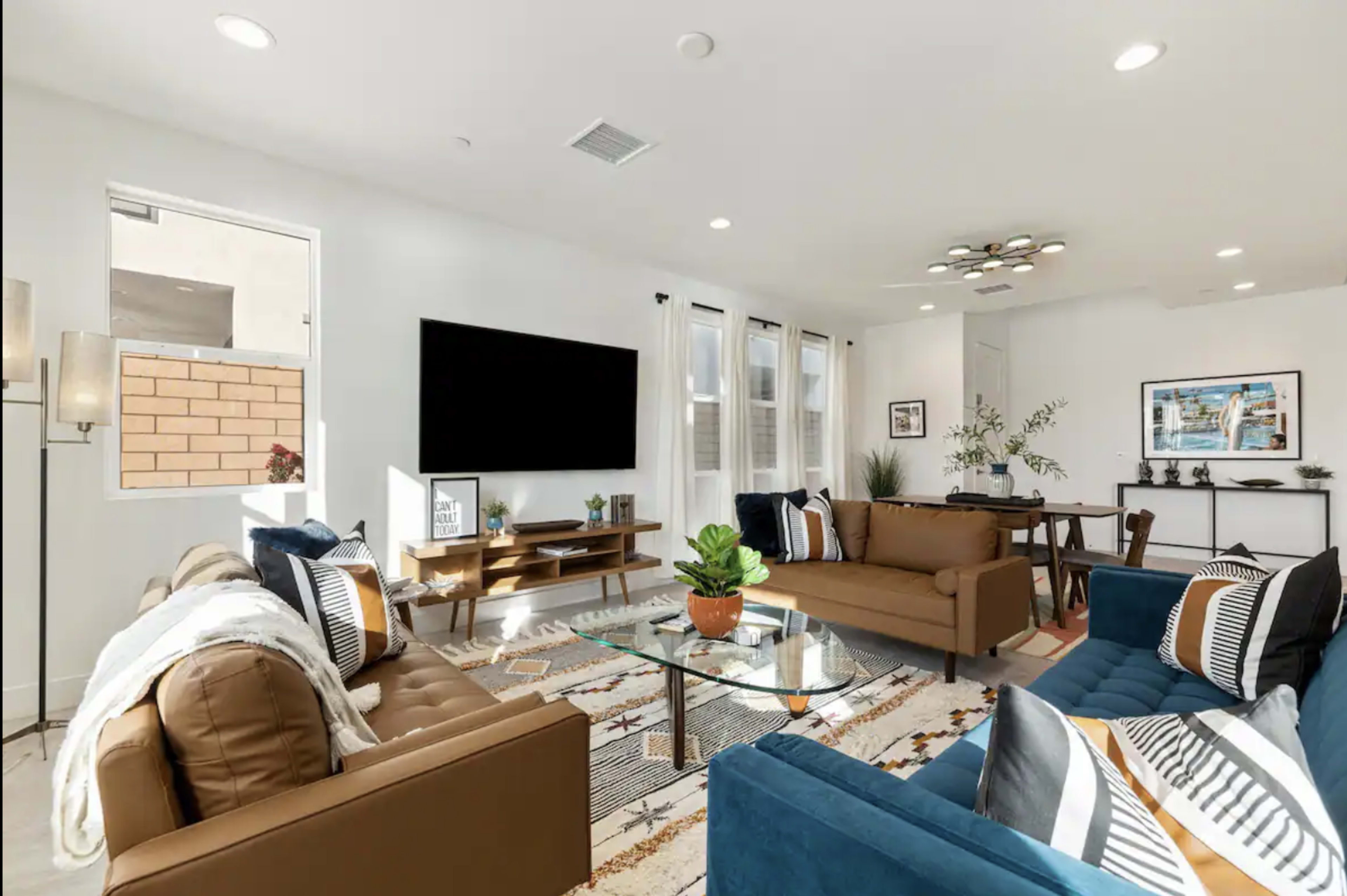 A modern living room with two sofas, a glass coffee table, a mounted television, and a dining area with a small table, all arranged around a patterned area rug.