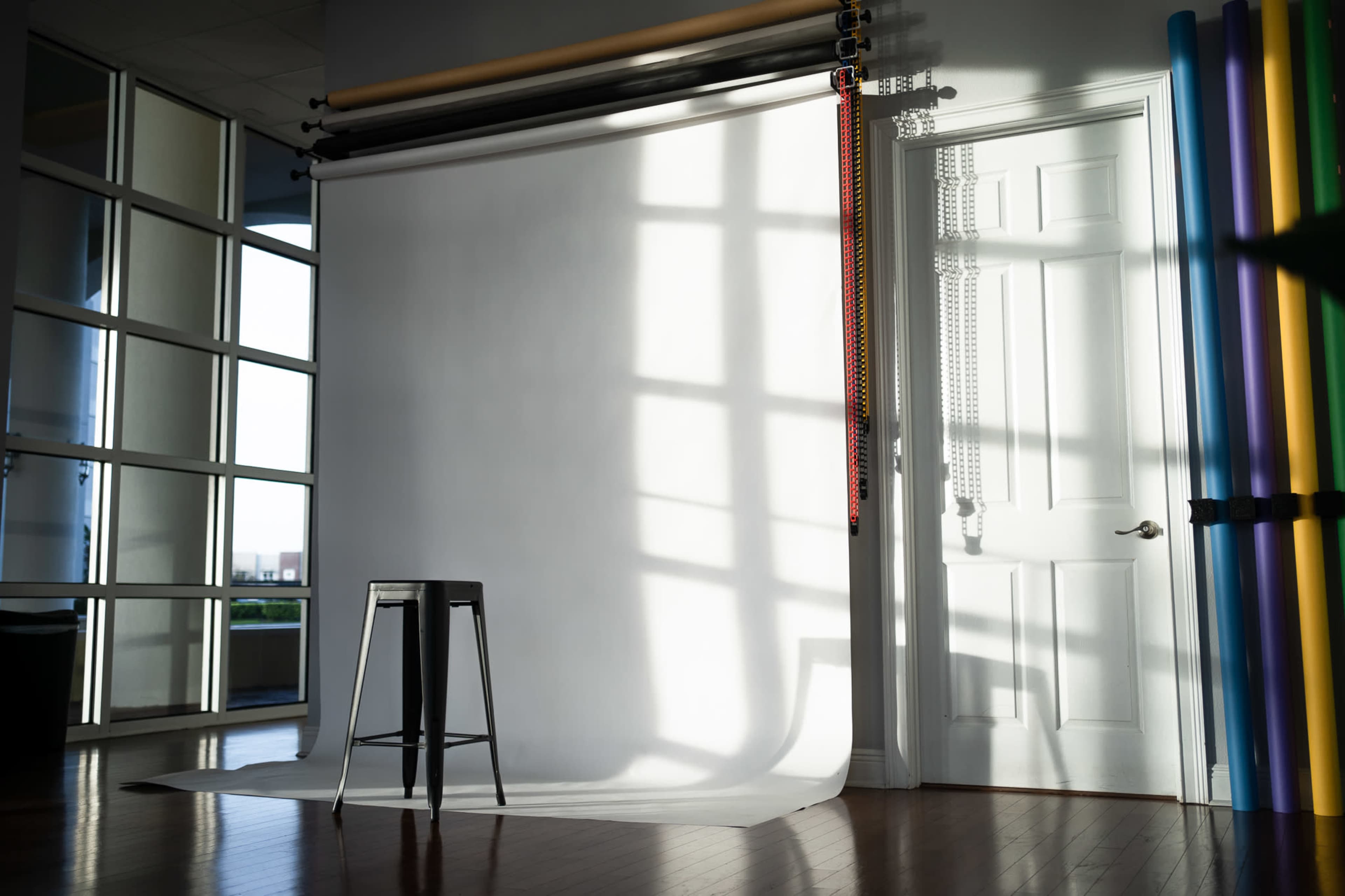 The image shows a photography studio with a blank white backdrop, a black stool, and colorful backdrops hanging on the wall.