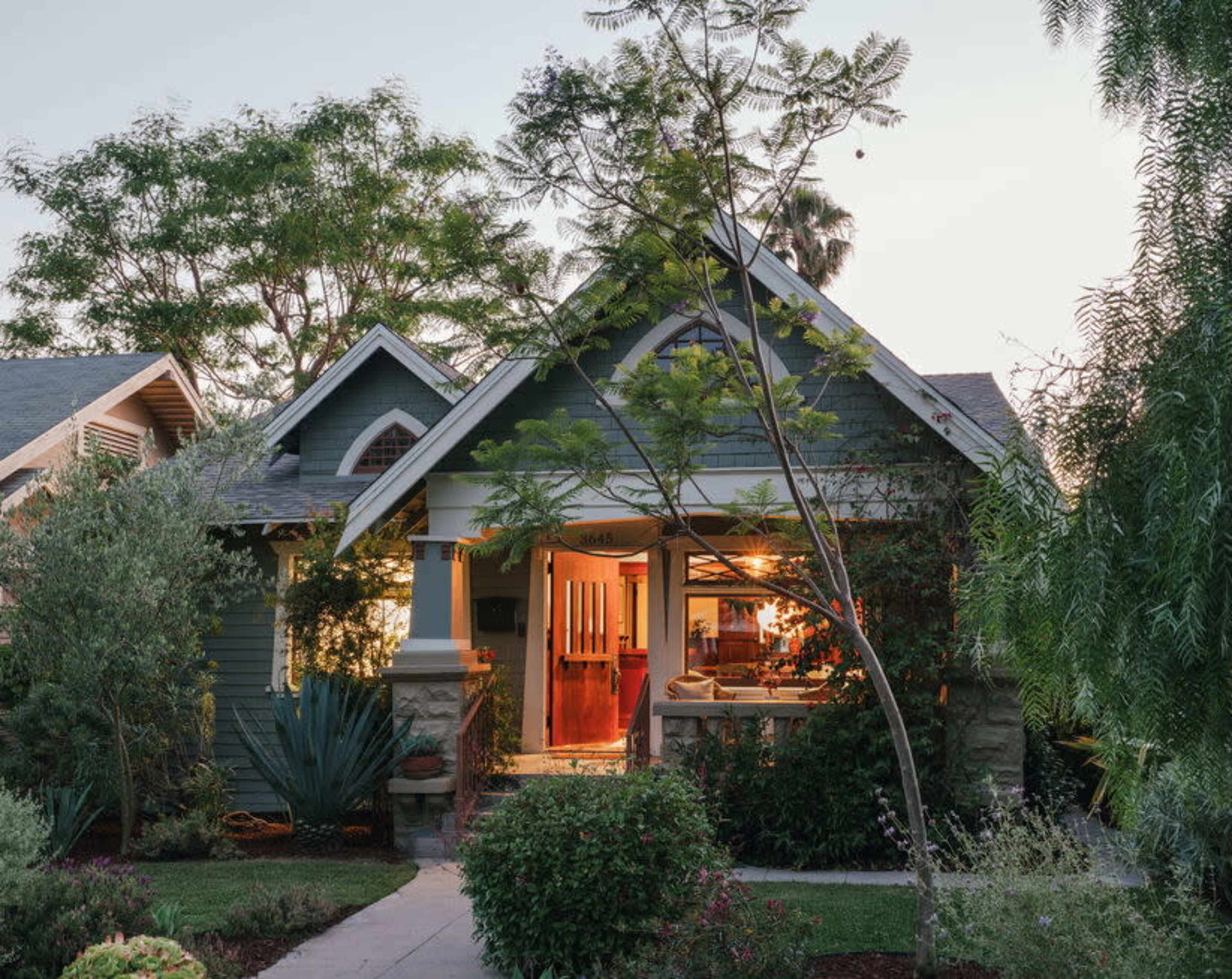 A craftsman-style house with a welcoming front porch is illuminated at dusk, surrounded by lush greenery and landscaping.