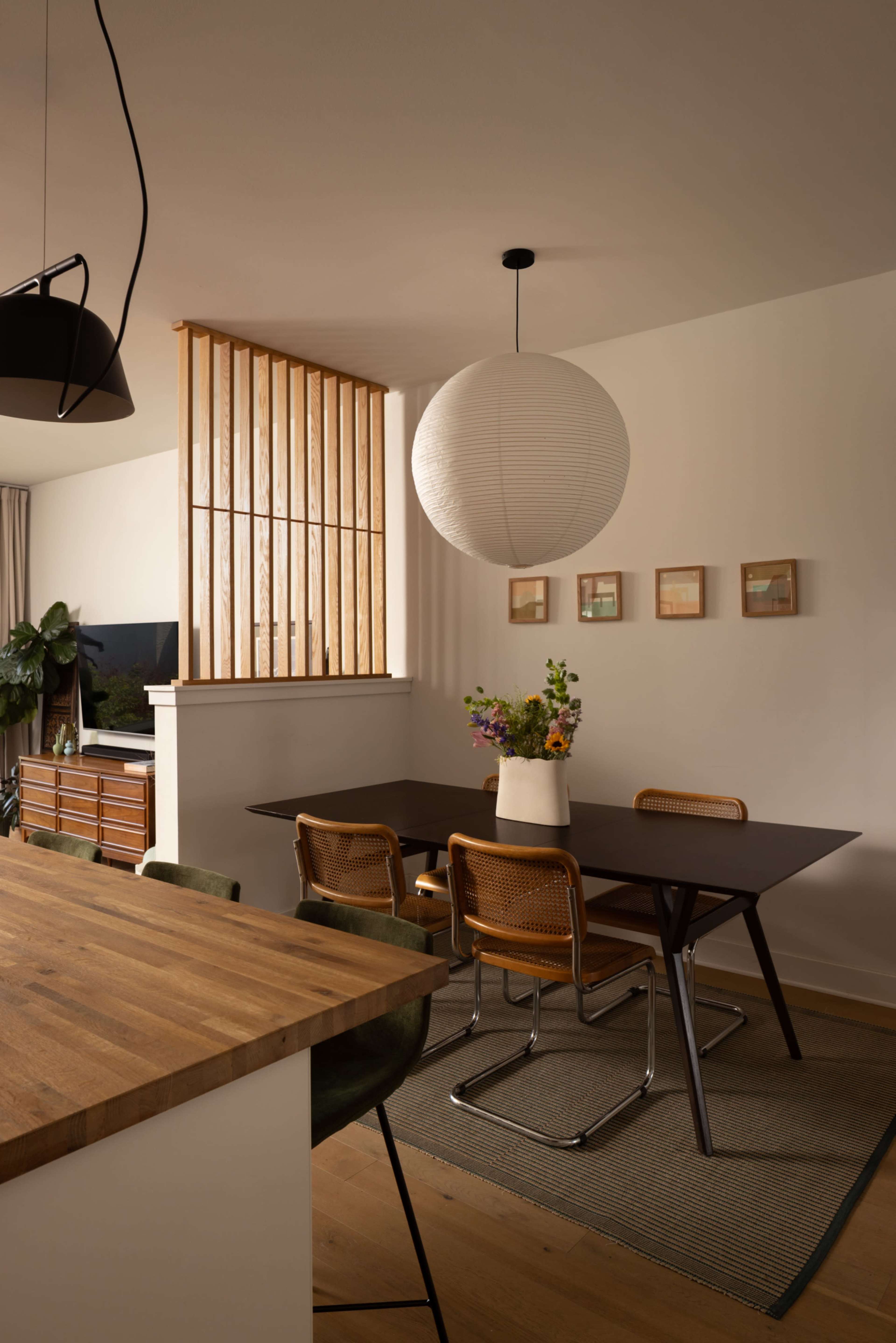 A modern dining area features a wooden table, four chairs, a large pendant light, and a decorative wall with framed pictures and wooden slats.