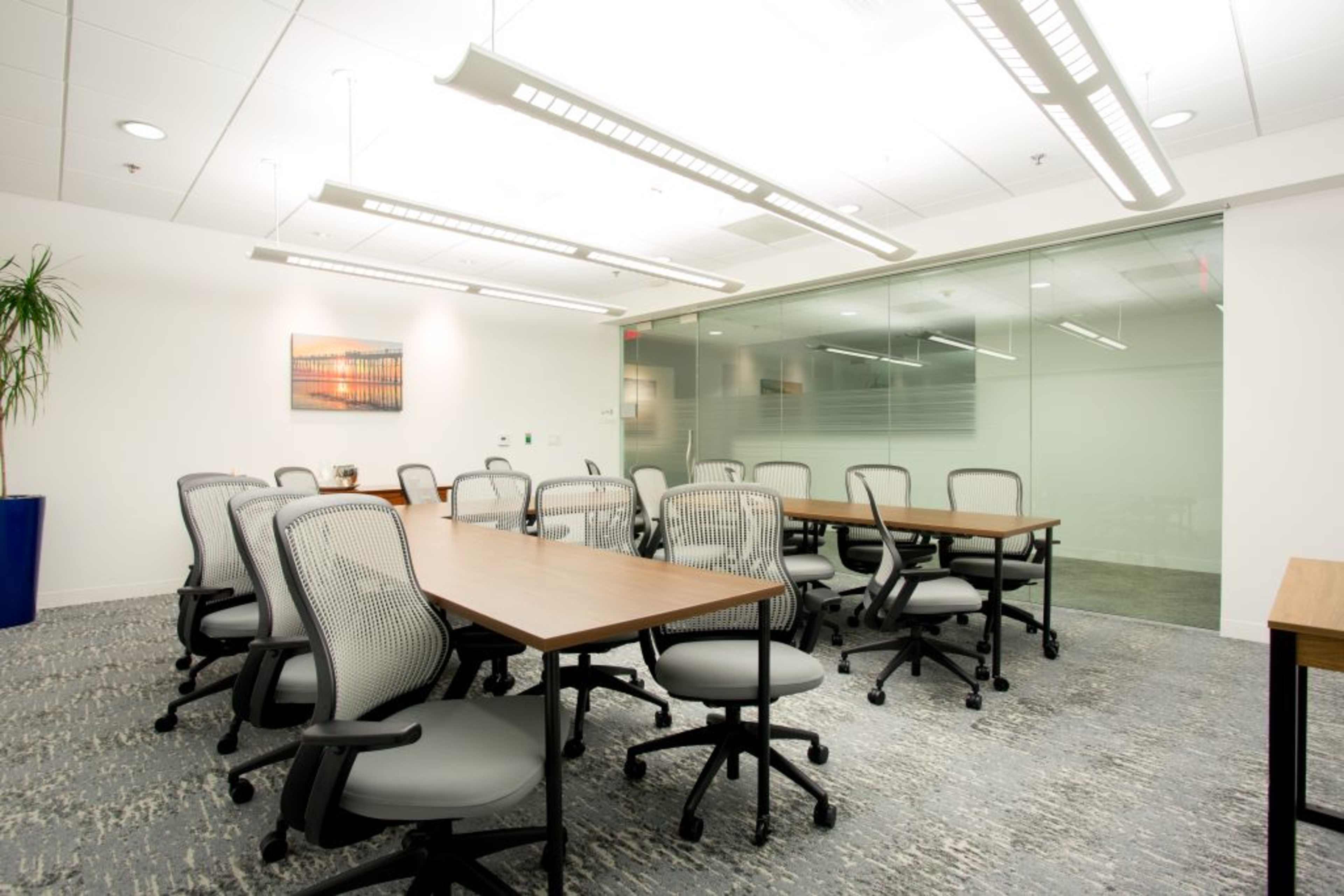 A modern meeting room features several tables and chairs arranged around a central space, with a plant and artwork on the walls.