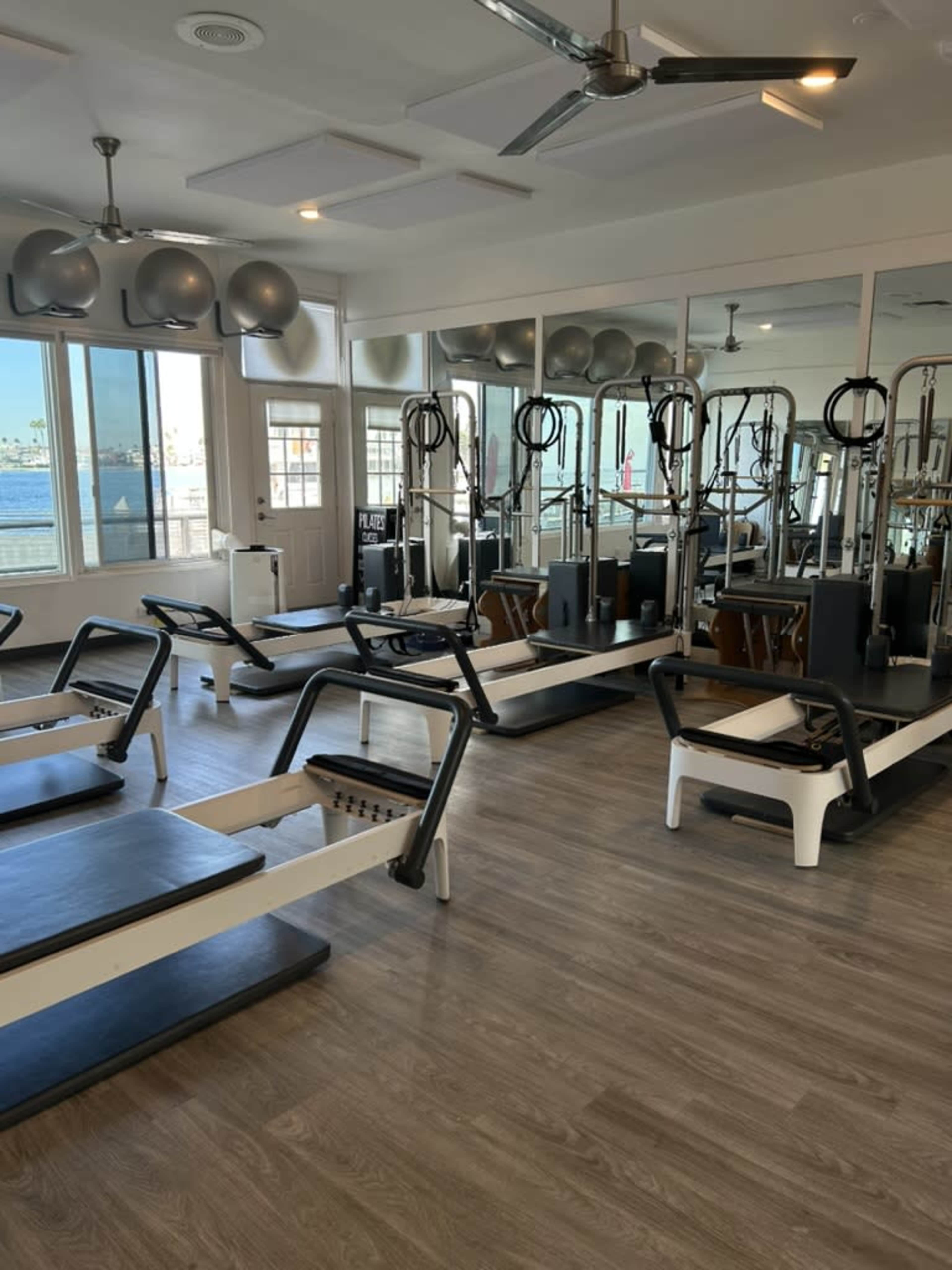 The image shows a spacious fitness studio equipped with multiple reformer machines, large mirrors, and a view of the waterfront through a window.