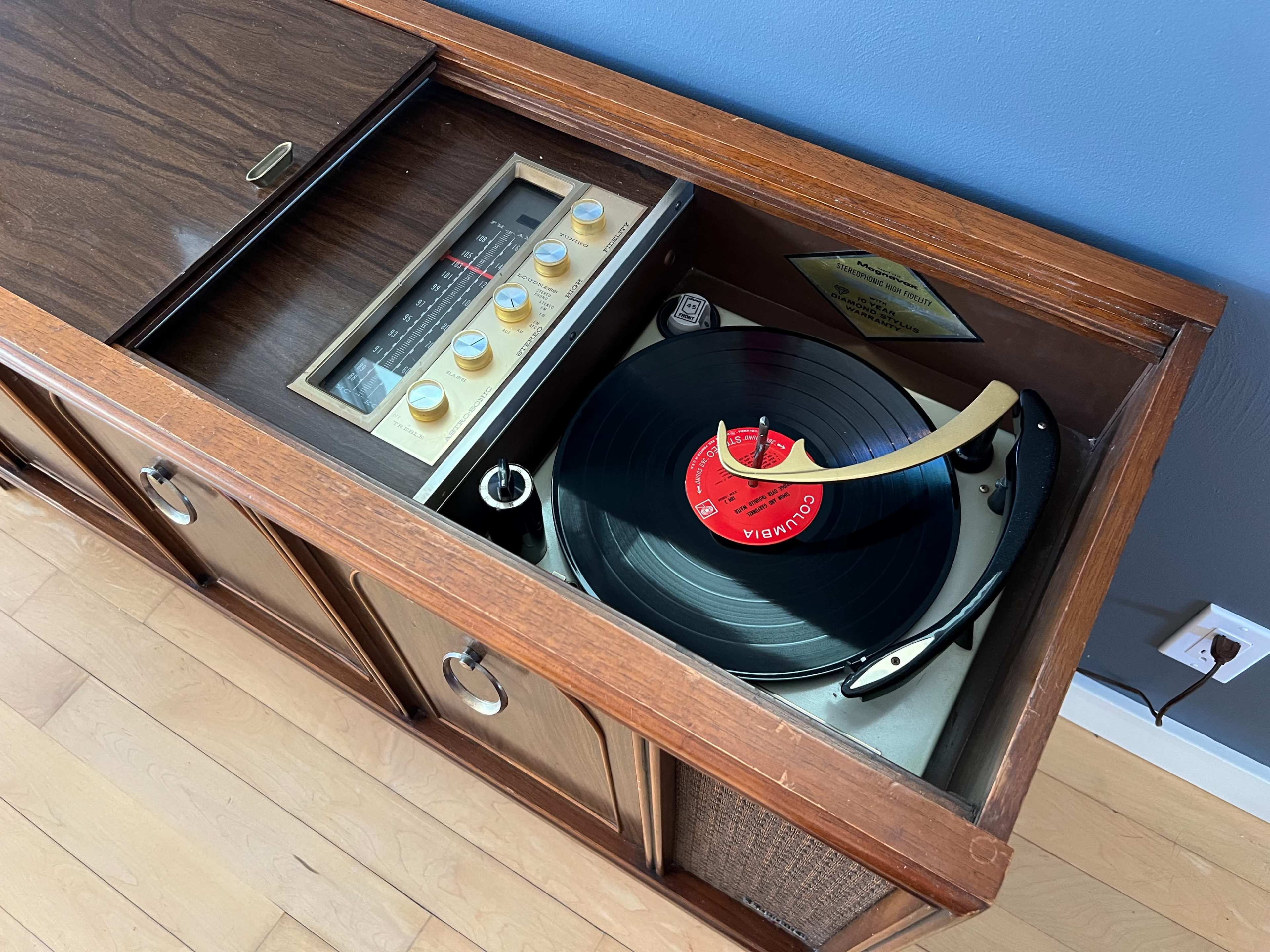 A vintage record player with a vinyl record is housed in a wooden cabinet with an integrated radio and control knobs.