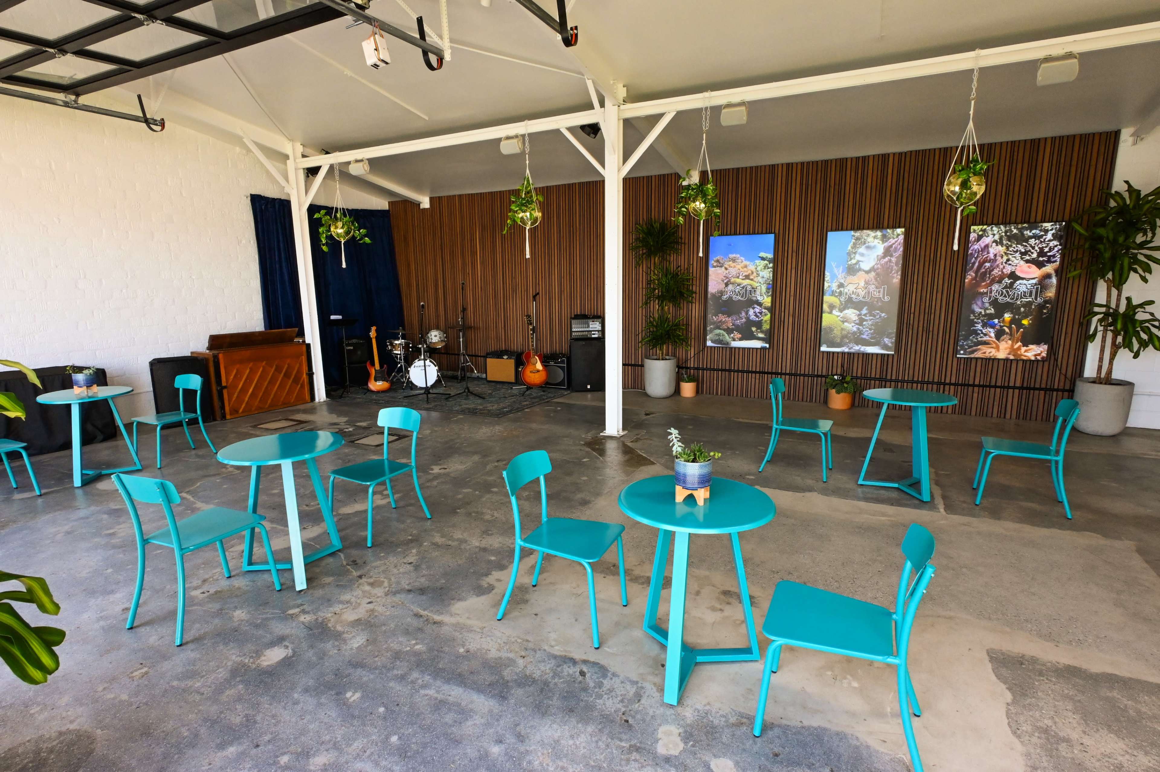 A spacious indoor area featuring turquoise tables and chairs, potted plants, and a small stage set up for musical instruments against a wooden backdrop.