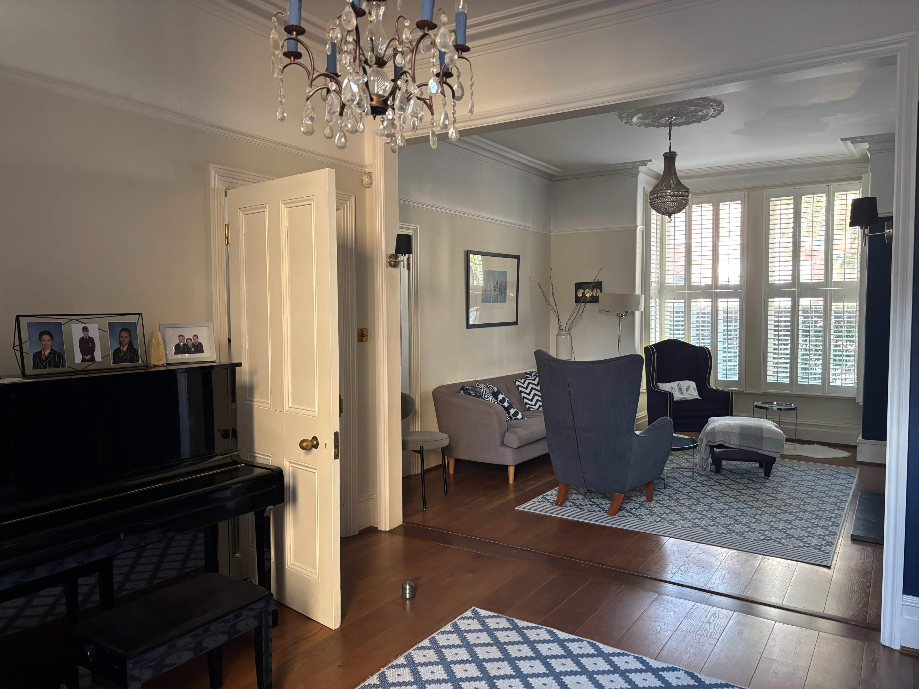 The image shows a living room with a piano, a couch, a chair, and large windows with shutters, along with a chandelier and patterned rugs on the wooden floor.