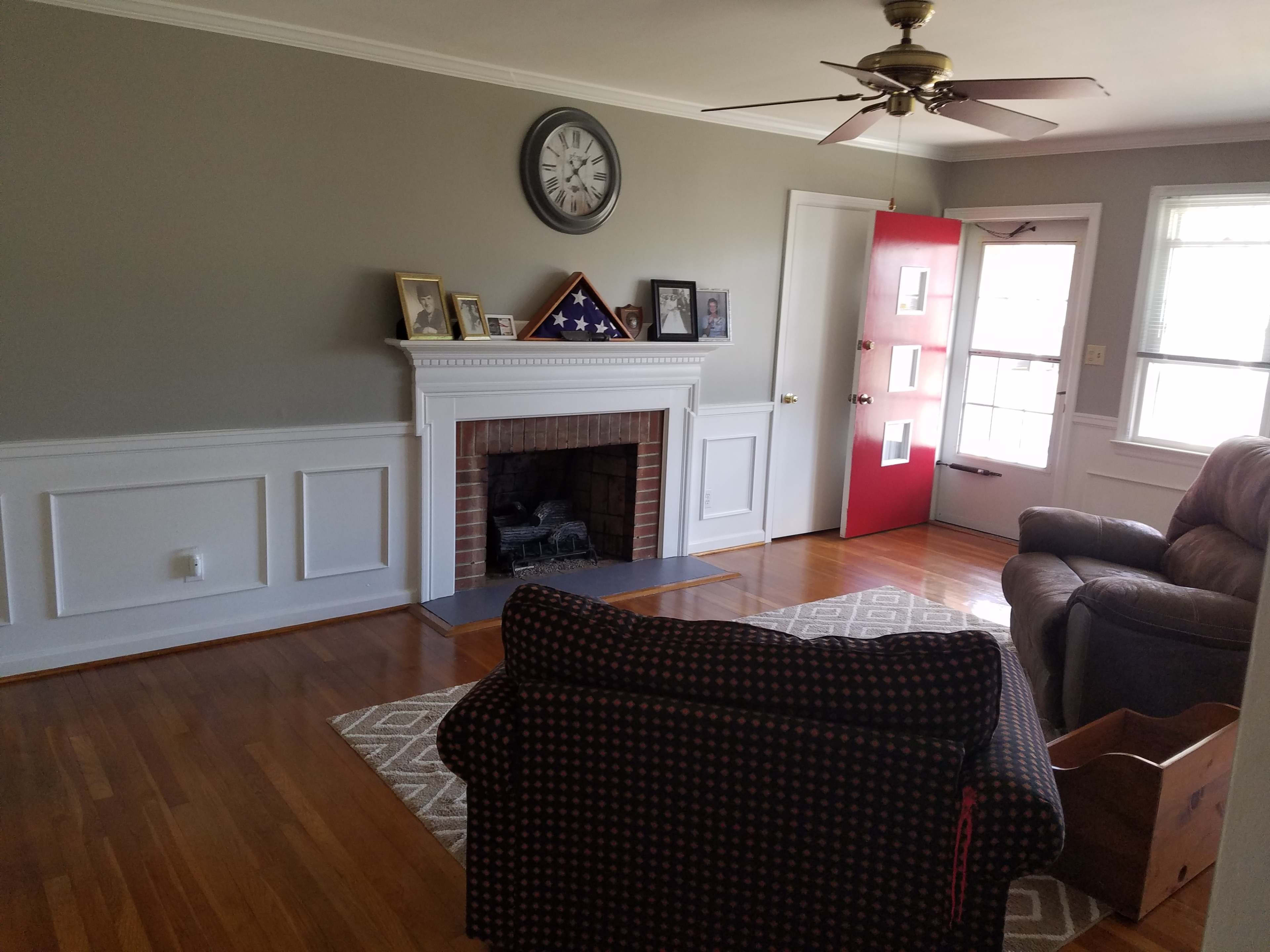 The living room features a fireplace with a white mantel, a large wall clock, framed pictures, a red door, and a brown armchair on a patterned rug.