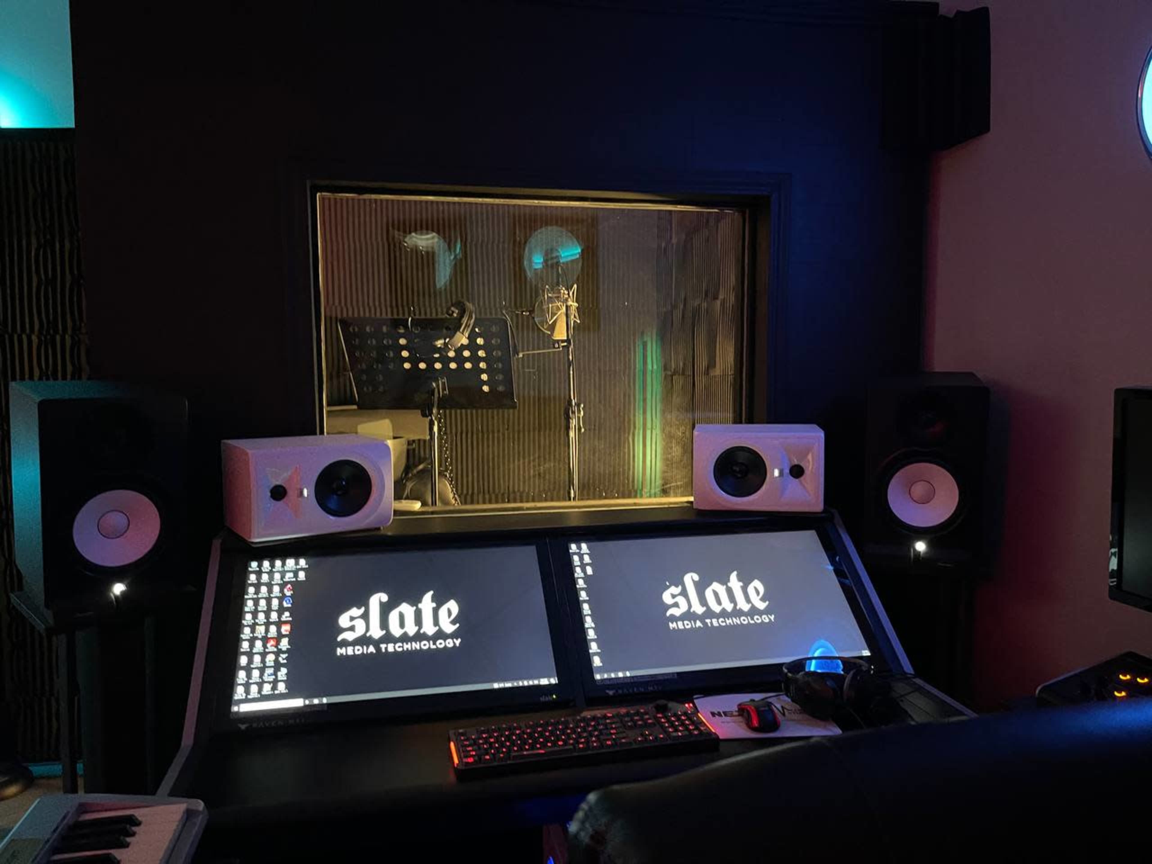 A recording studio with dual computer monitors displaying "slate MEDIA TECHNOLOGY" and a sound booth in the background.