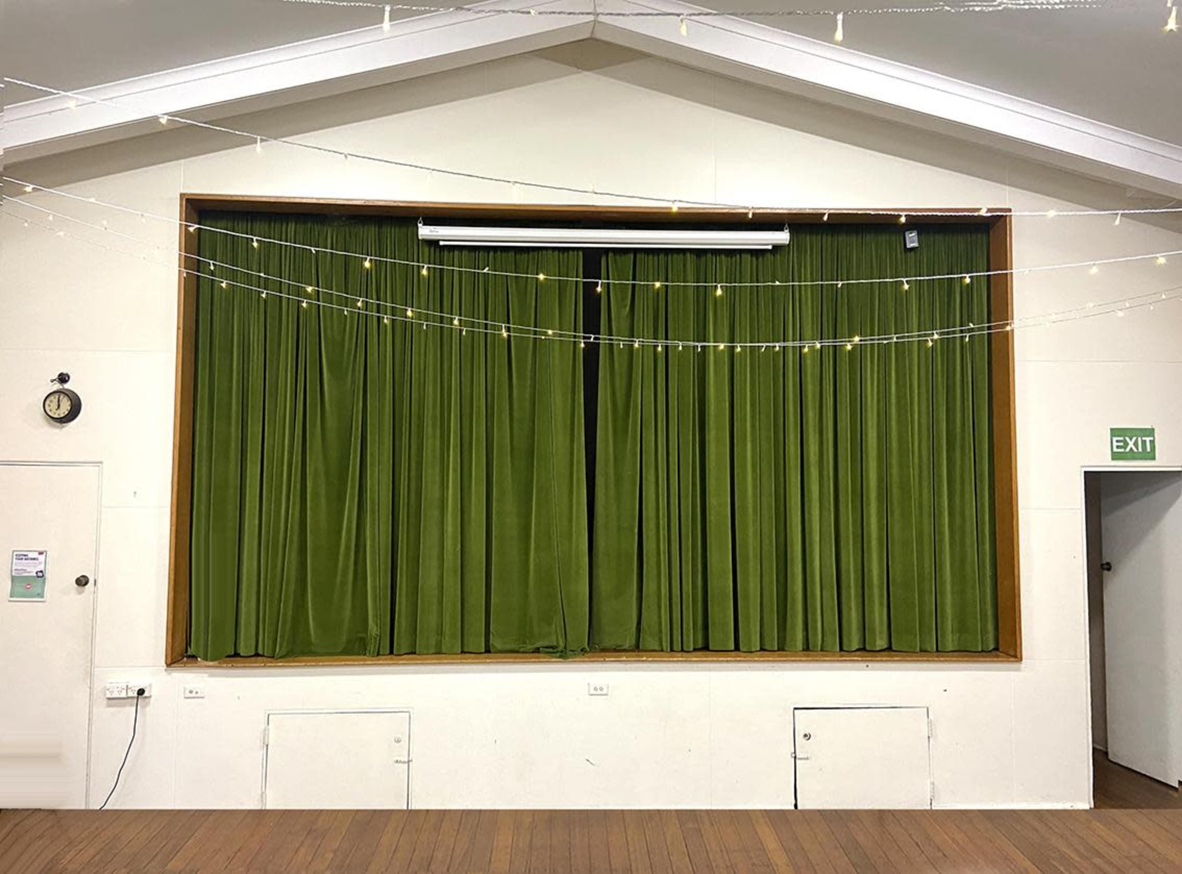 The image shows a stage with closed green curtains in a room featuring wooden flooring and string lights above.