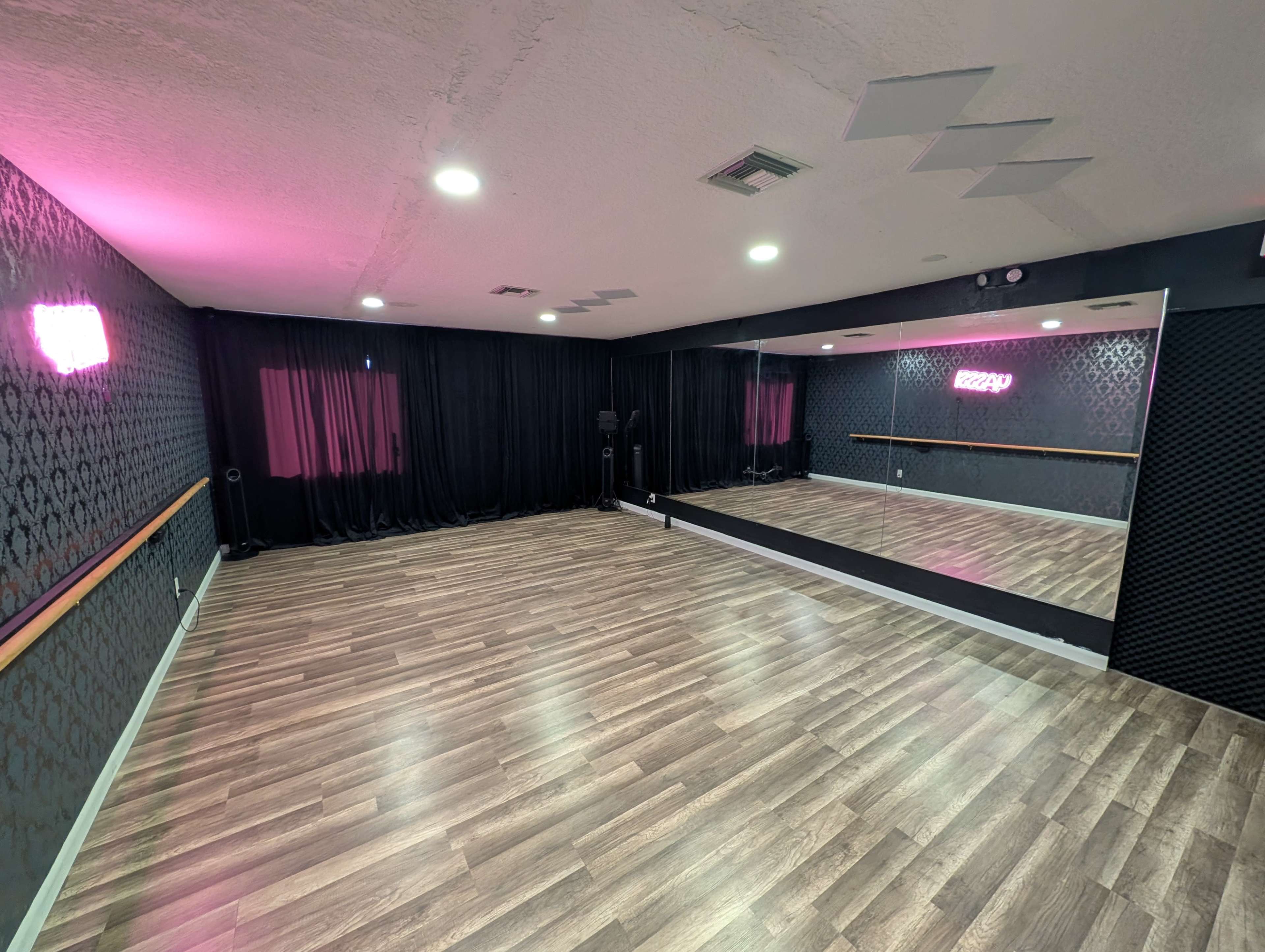 The image shows a dance studio featuring polished wooden floors, a large mirror on one wall, and black curtains along another wall, illuminated by pink lighting.