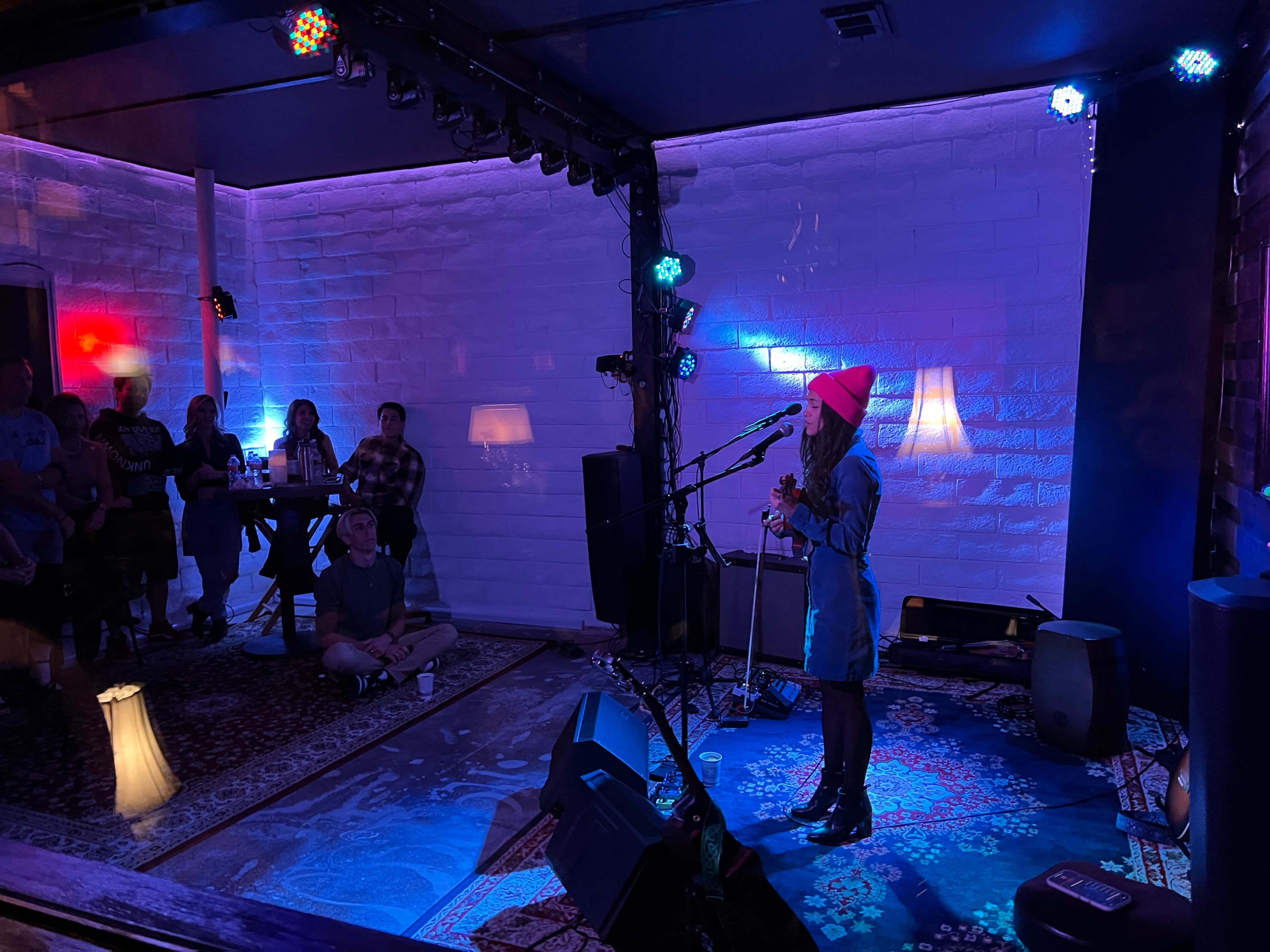 A performer stands on stage with a microphone in front of an audience seated in a dimly lit venue adorned with colorful lights.