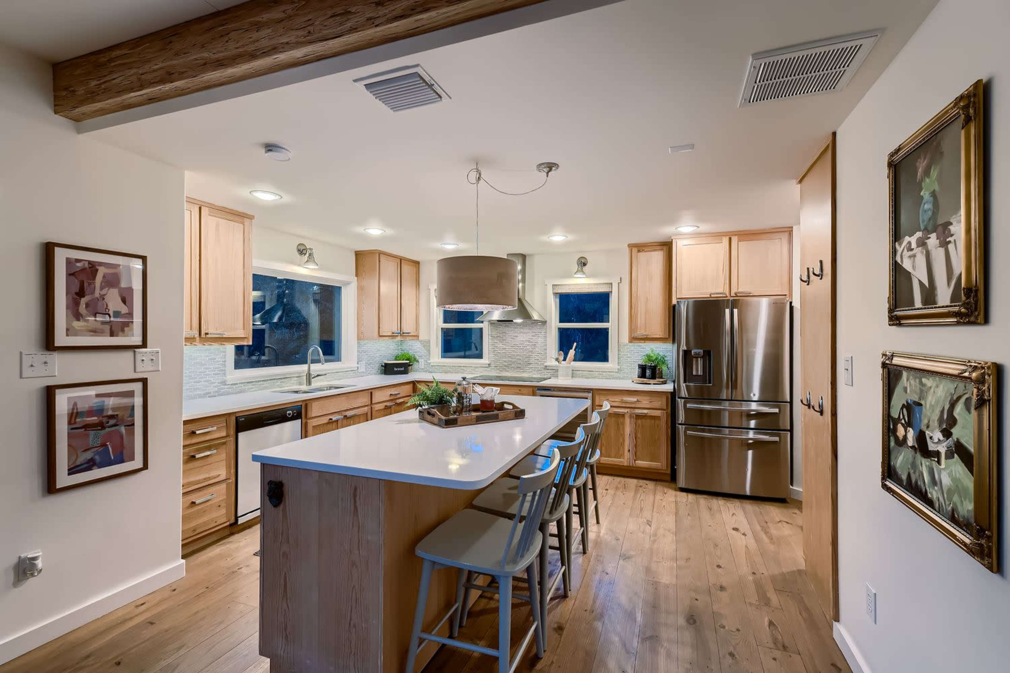 The image shows a modern kitchen with wooden cabinets, a large island with seating, and framed artwork on the walls.