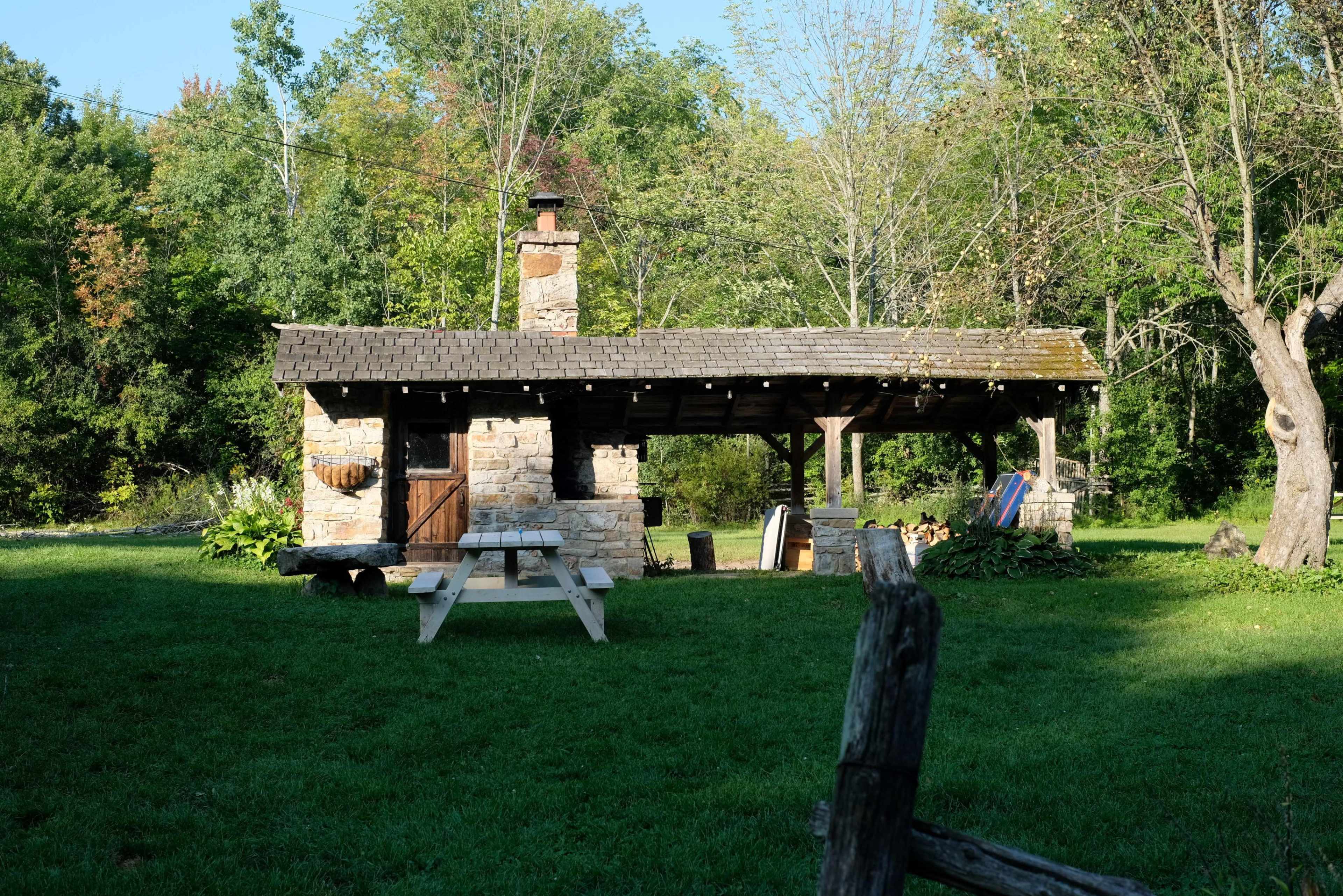 A stone cabin with a wooden roof and a chimney sits in a grassy area, surrounded by trees.