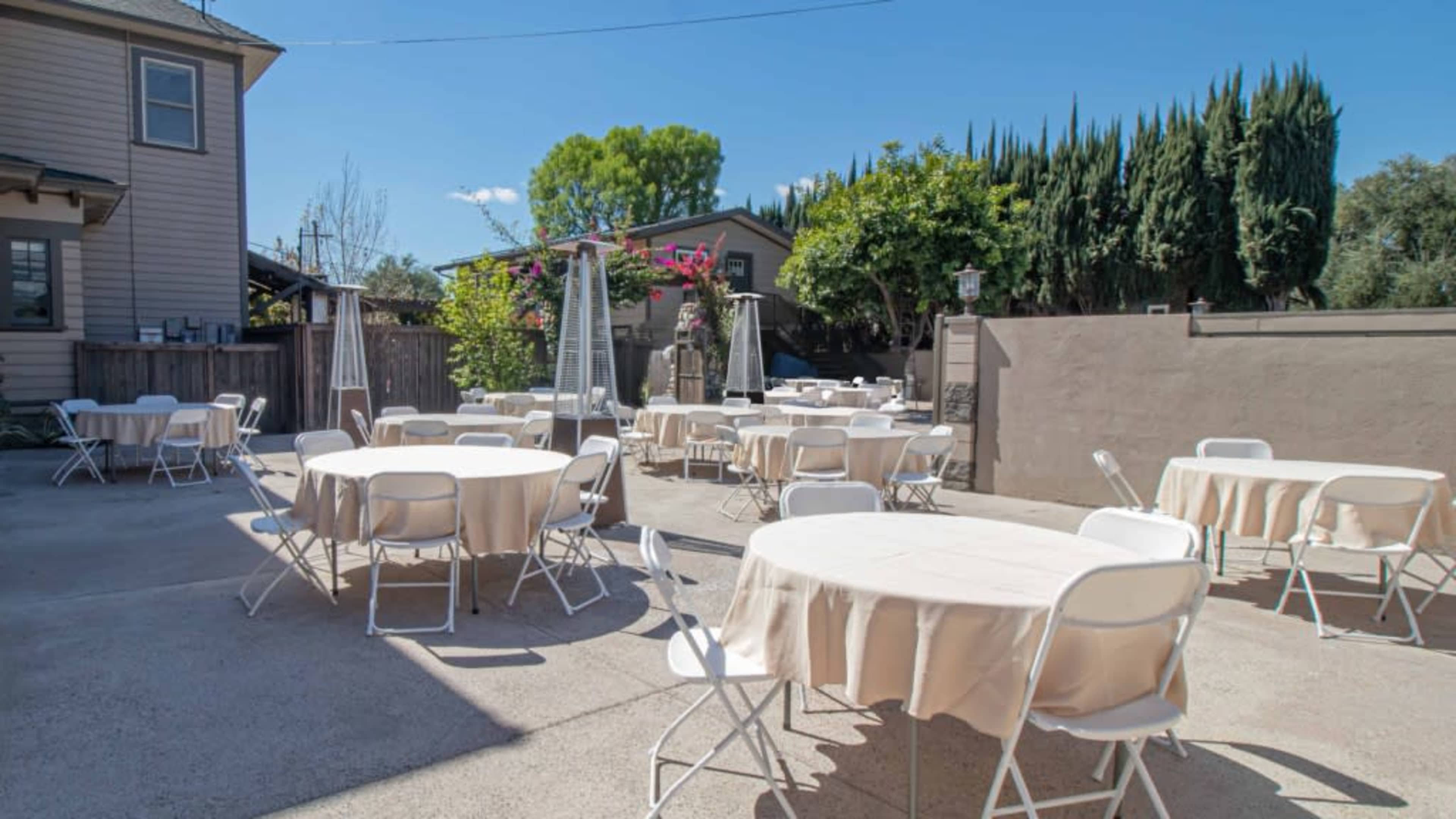 The image shows an outdoor seating area with several round tables covered with beige tablecloths, surrounded by a fenced space and greenery.