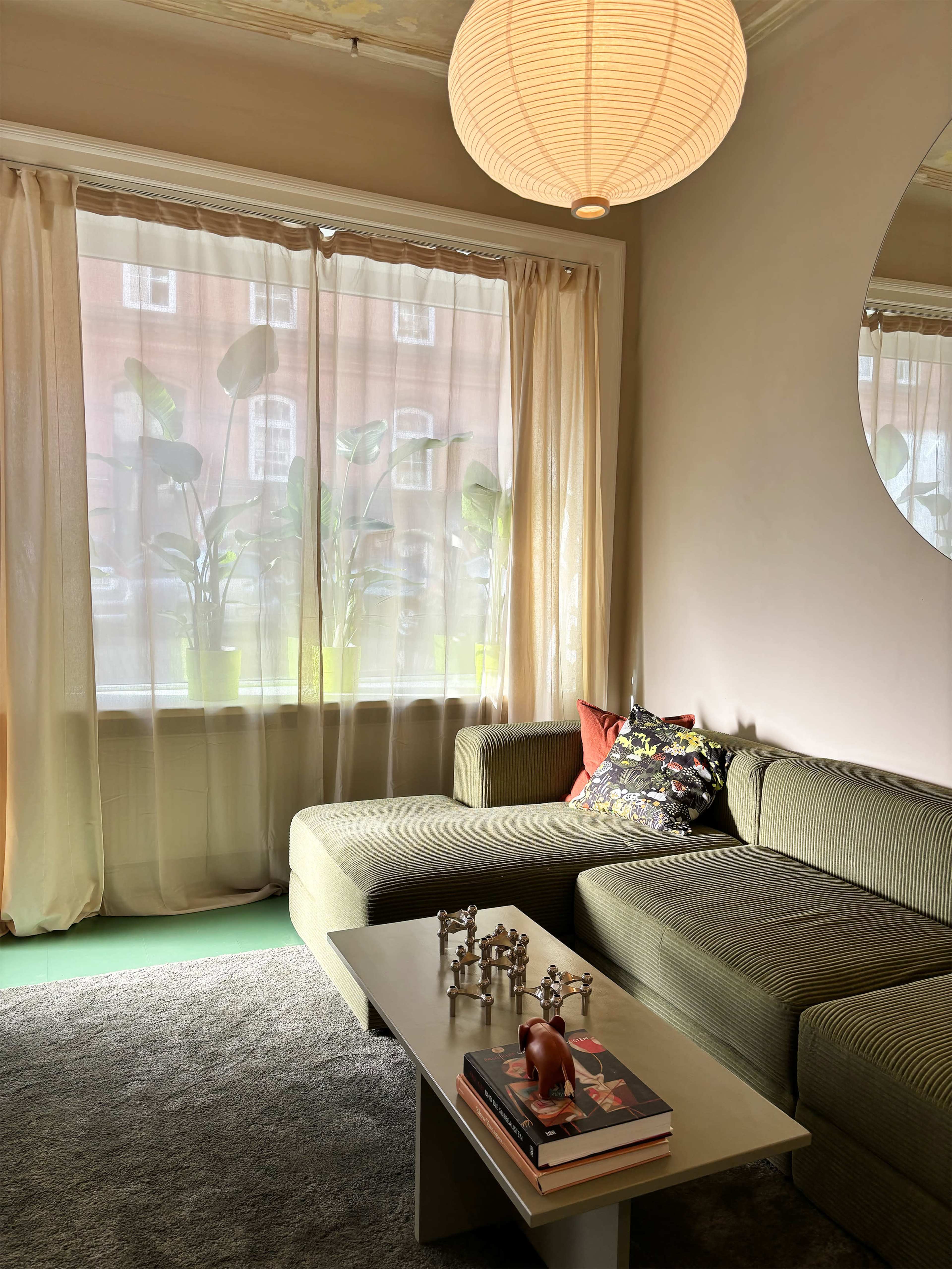 The image shows a modern living room with a green velvet sofa, a coffee table with decorative items, and large windows covered by sheer curtains.