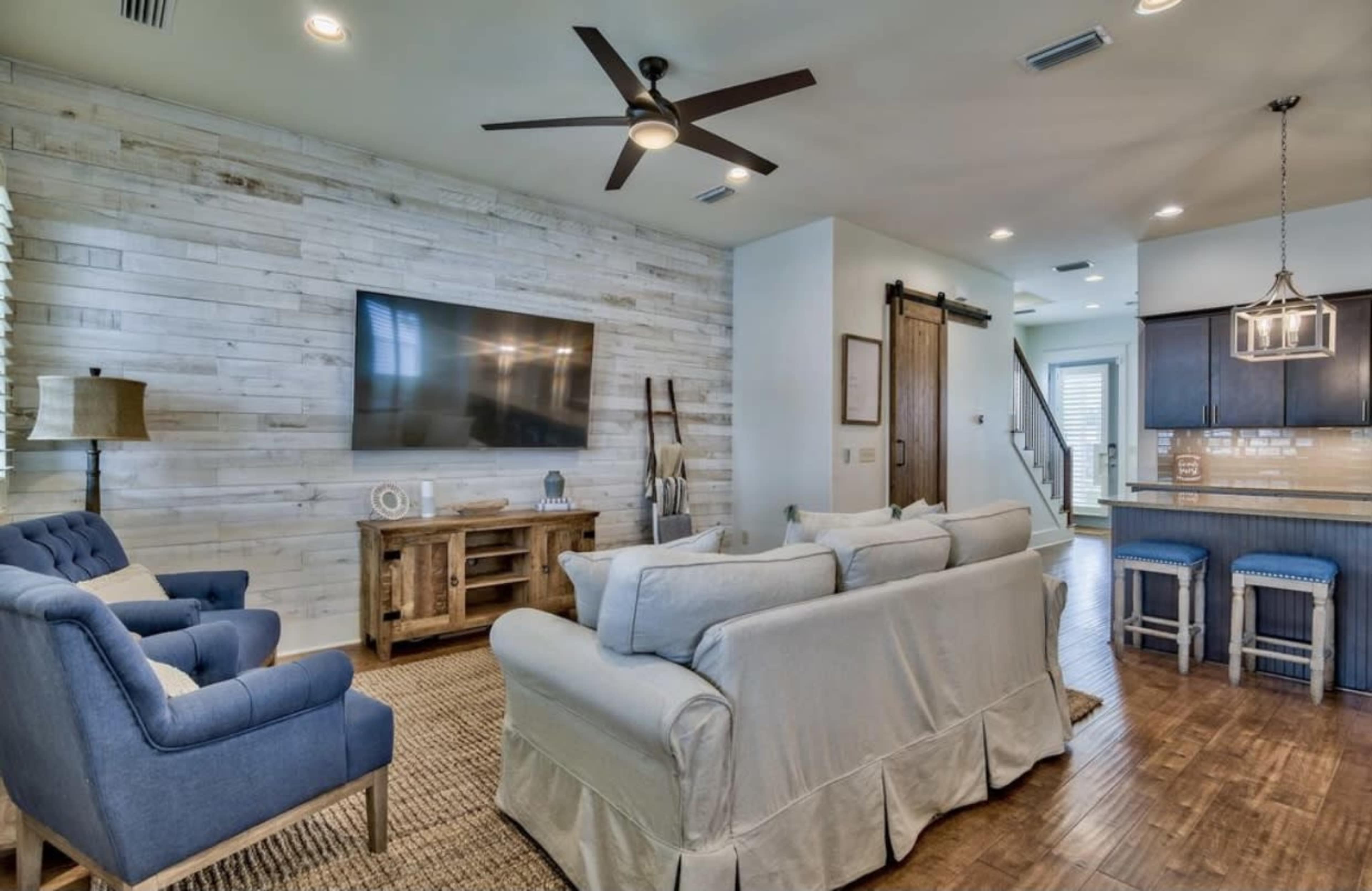 A modern living room with light-colored walls, a large flat-screen TV, wooden flooring, and a seating area featuring a blue armchair and a light-colored sofa.