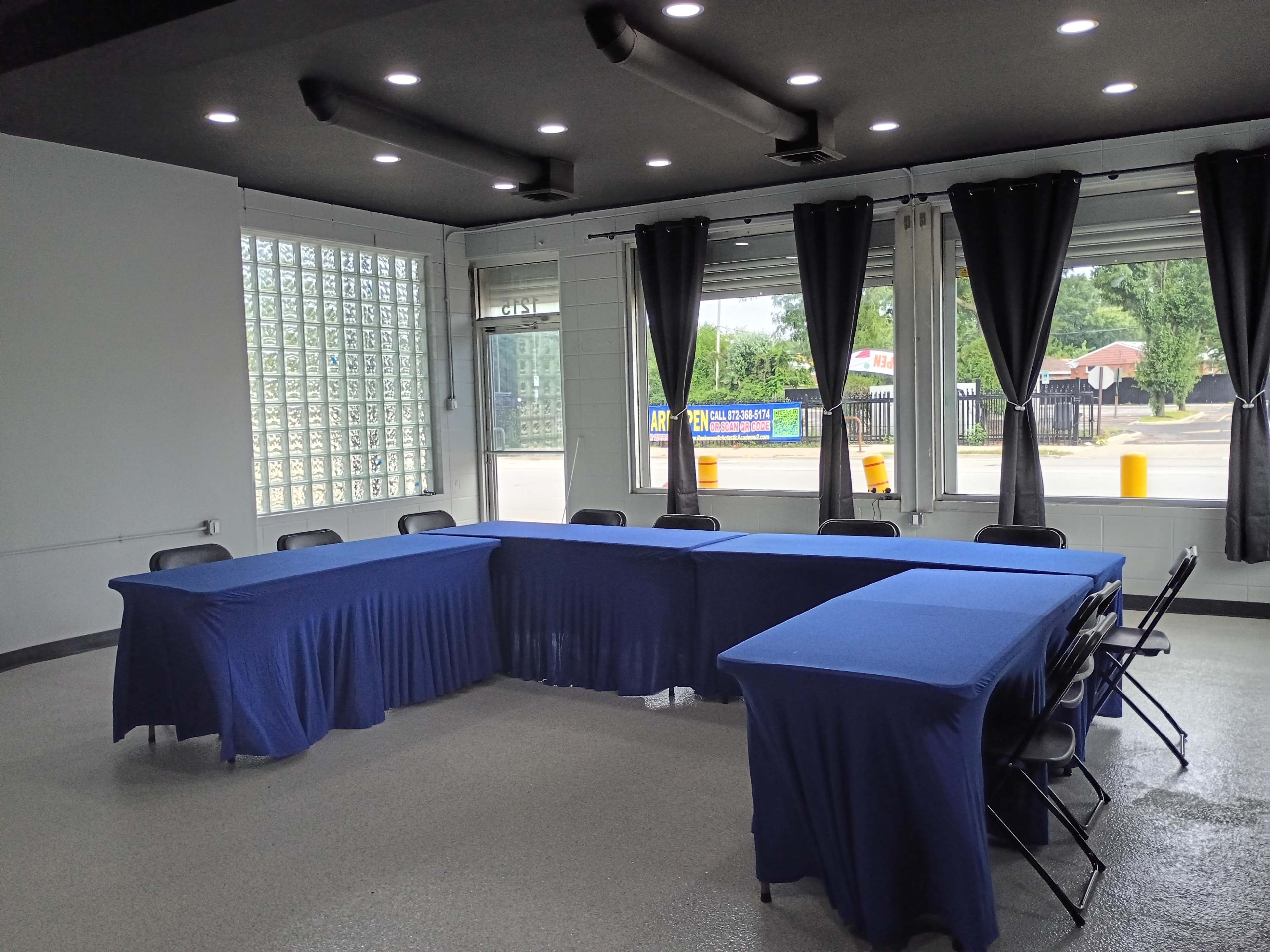 A conference room is set up with blue tablecloths covering several tables arranged in a U-shape, and large windows allow natural light to enter the space.