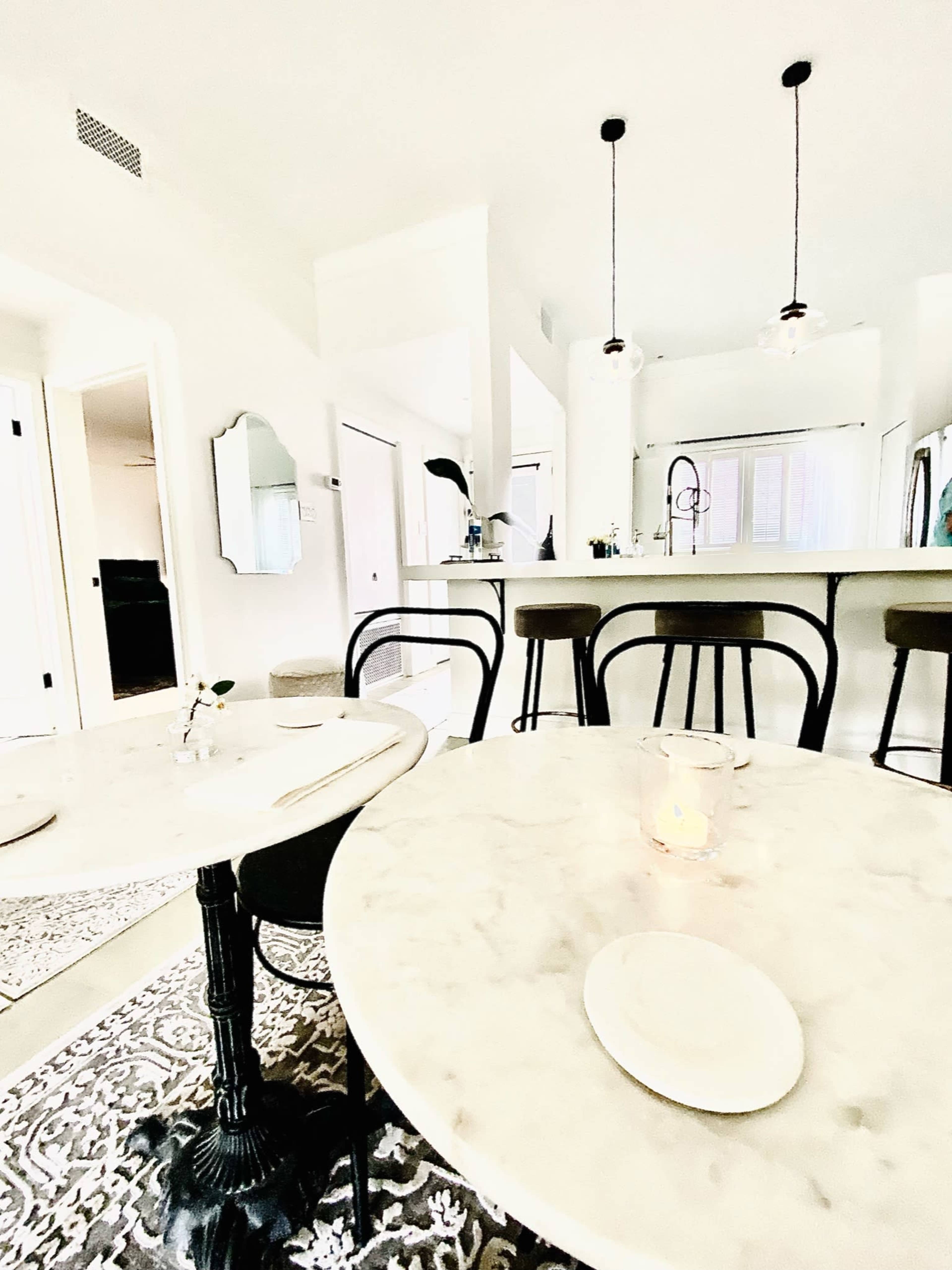 A bright restaurant interior featuring marble-topped tables, black chairs, and a kitchen visible in the background.