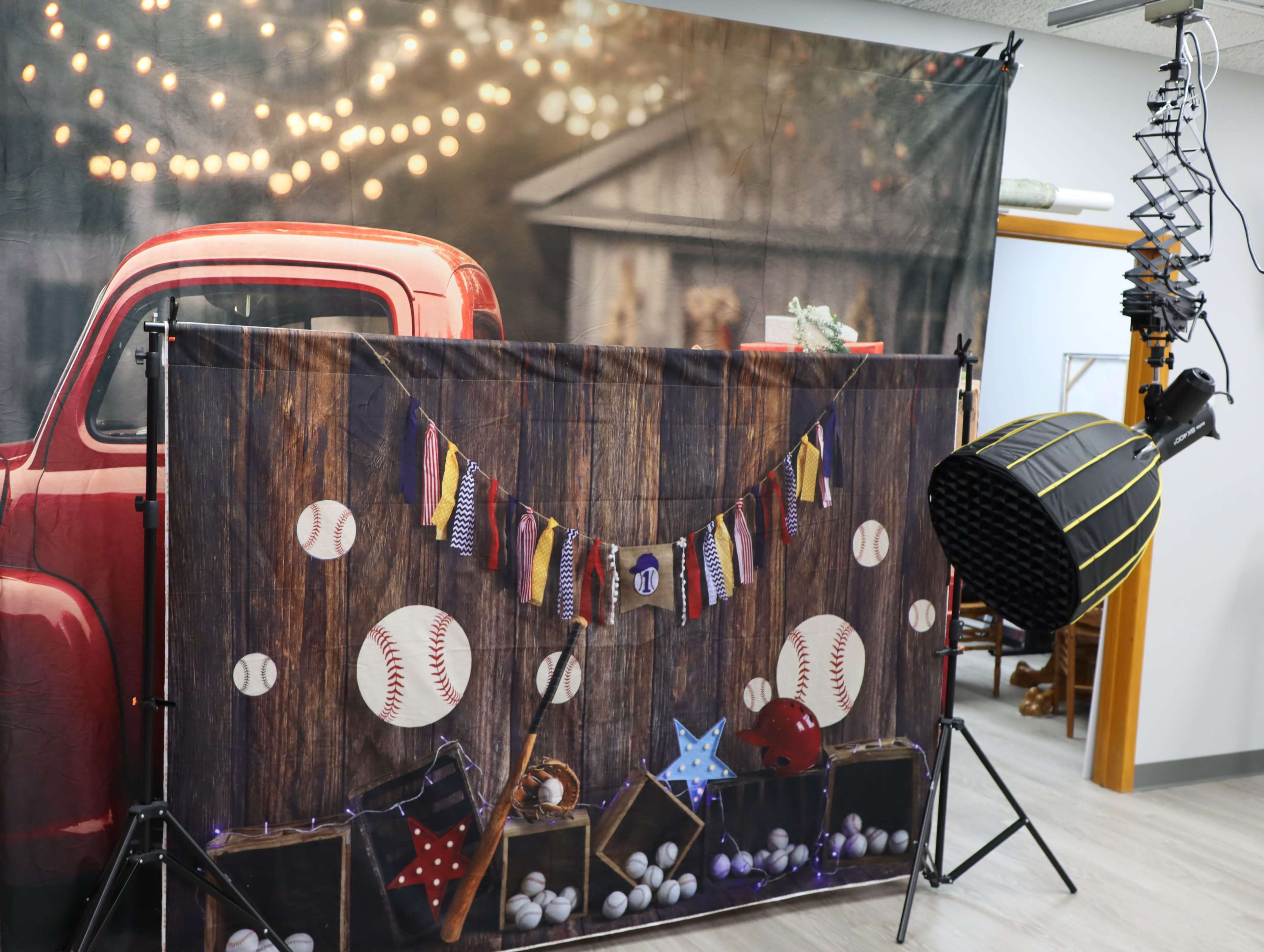 The image shows a photography setup featuring a vintage red truck backdrop and a decorated wooden panel with baseballs and star decorations.