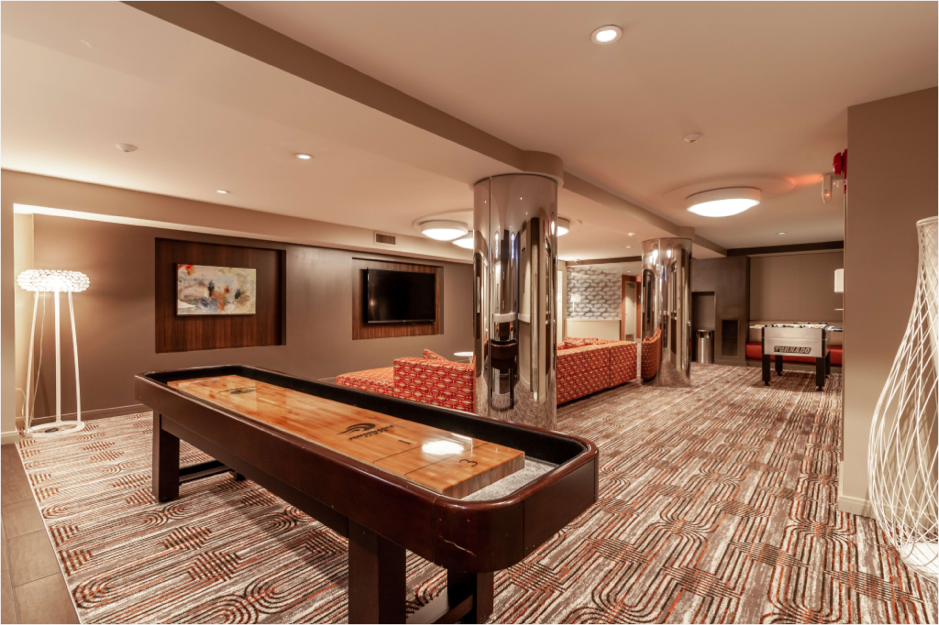 The image shows a modern lounge area featuring a shuffleboard table, cozy seating, and decorative lighting.