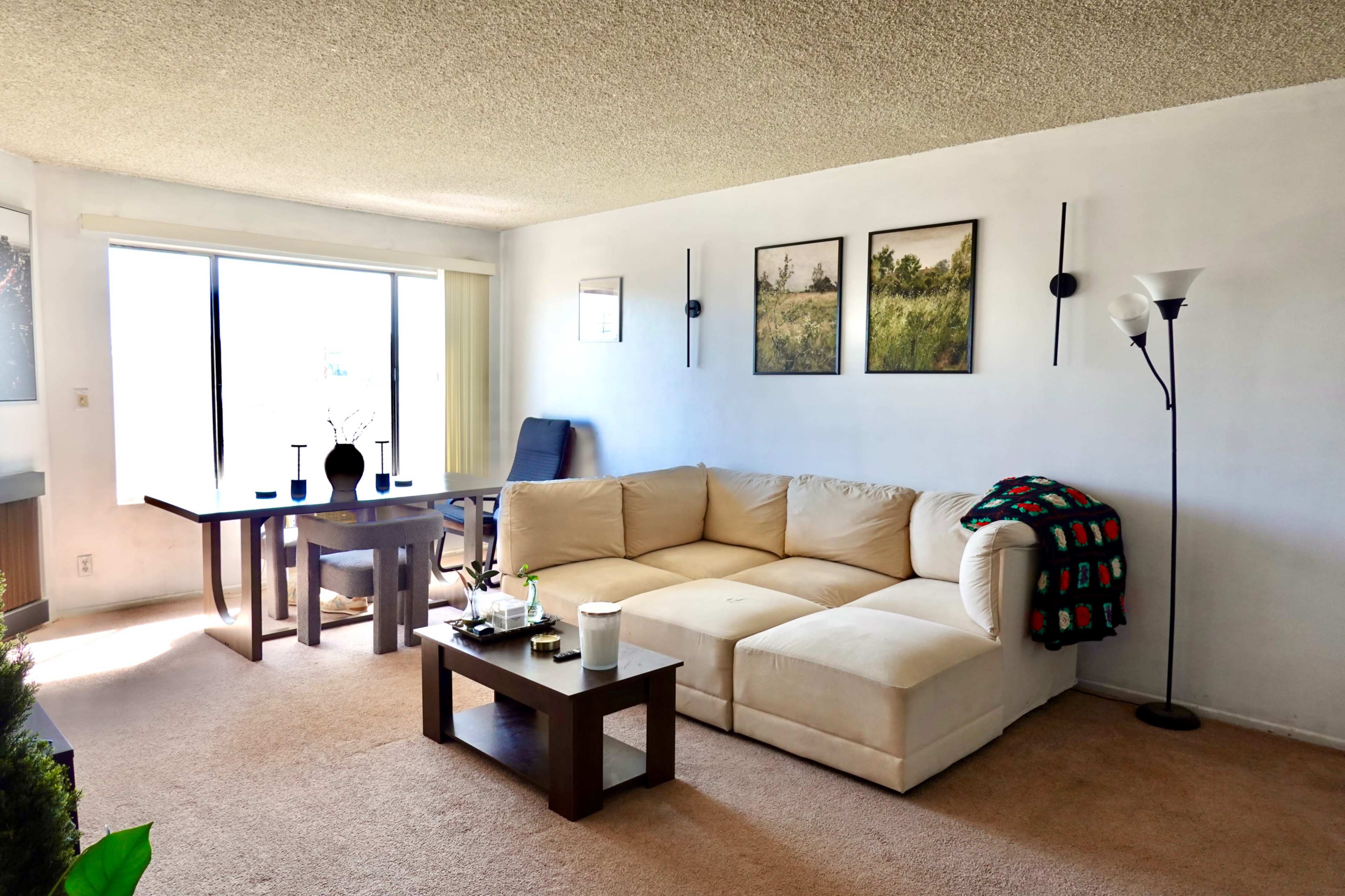 The image shows a living room with a beige sectional sofa, a small wooden coffee table, a dining table with chairs, and framed artwork on the wall.