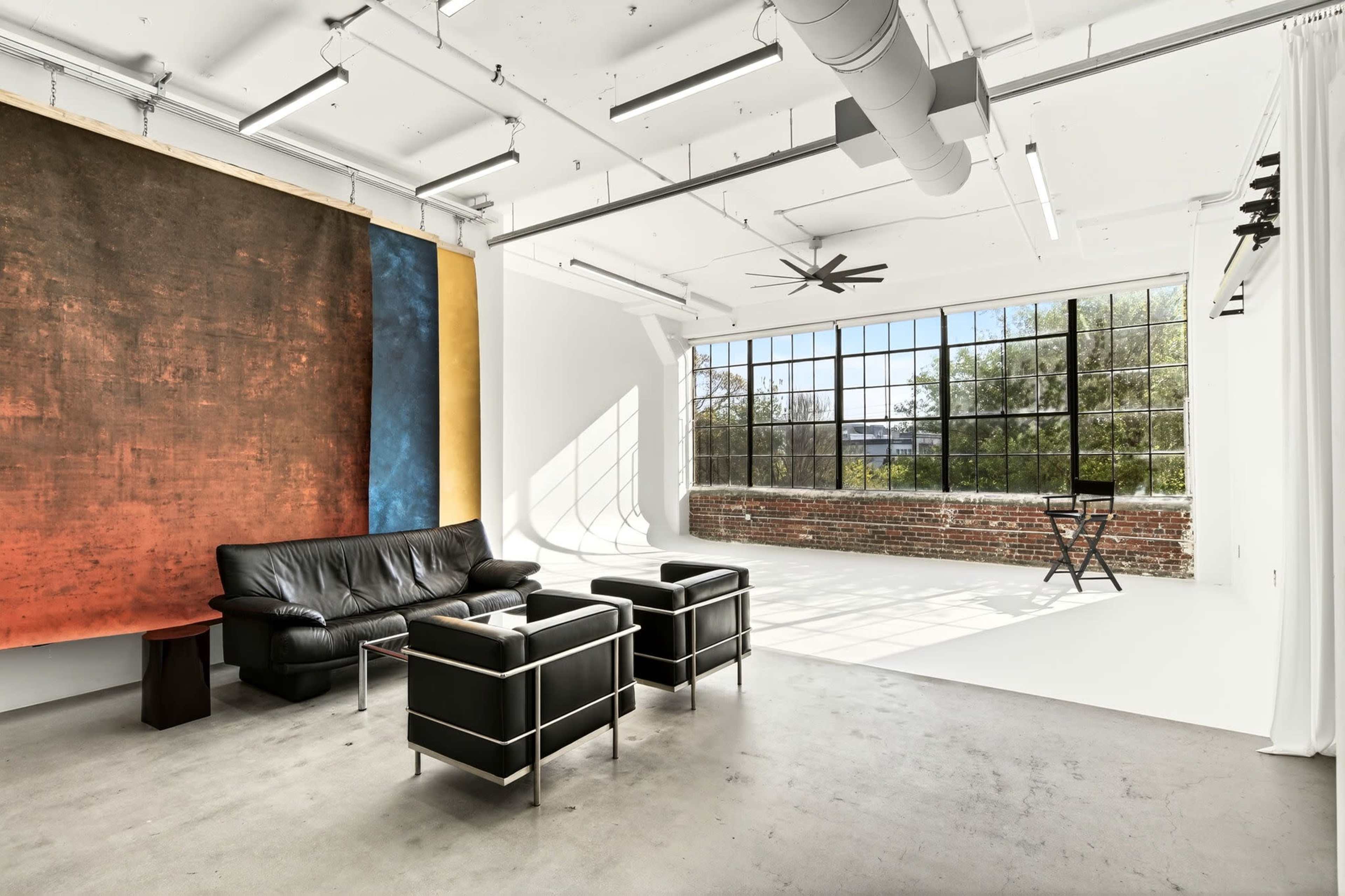 The image shows a modern, spacious interior with a black leather sofa, two black chairs, and a large window illuminating the room with natural light.