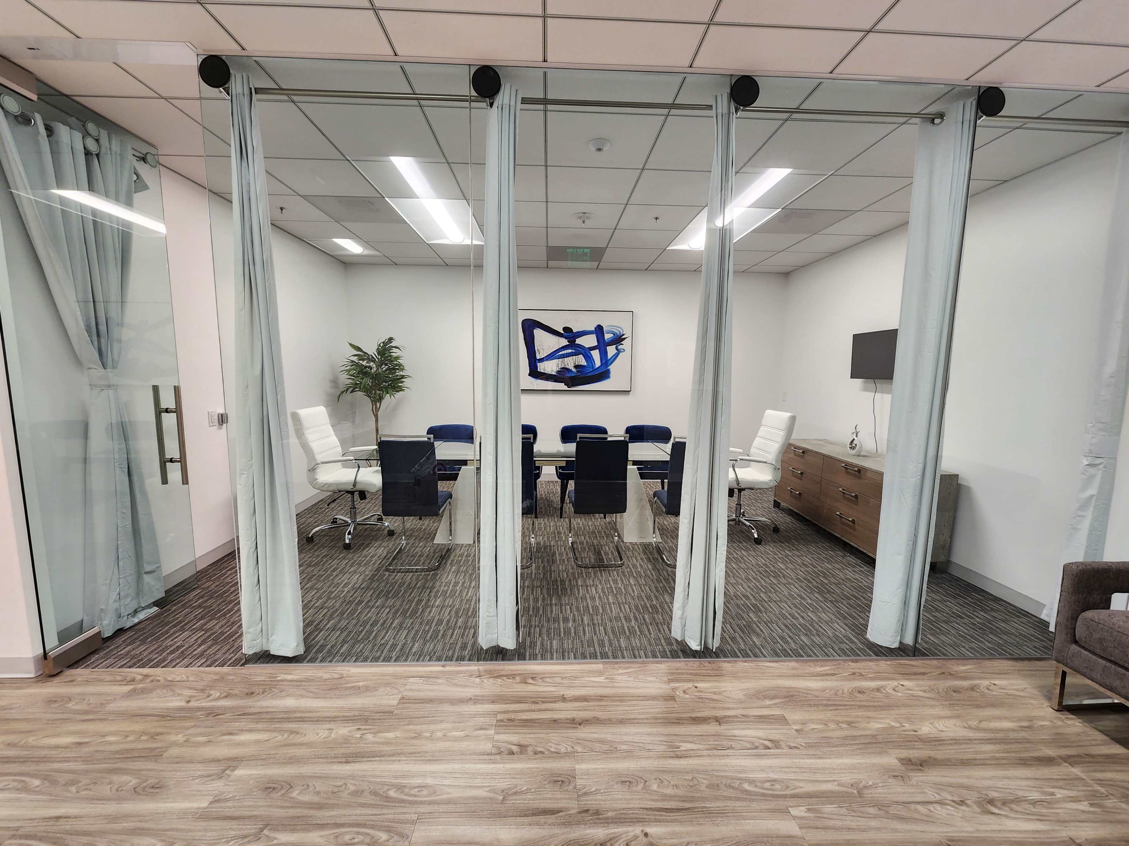 A glass-walled conference room features a large table surrounded by chairs, with a sideboard and artwork on the wall.