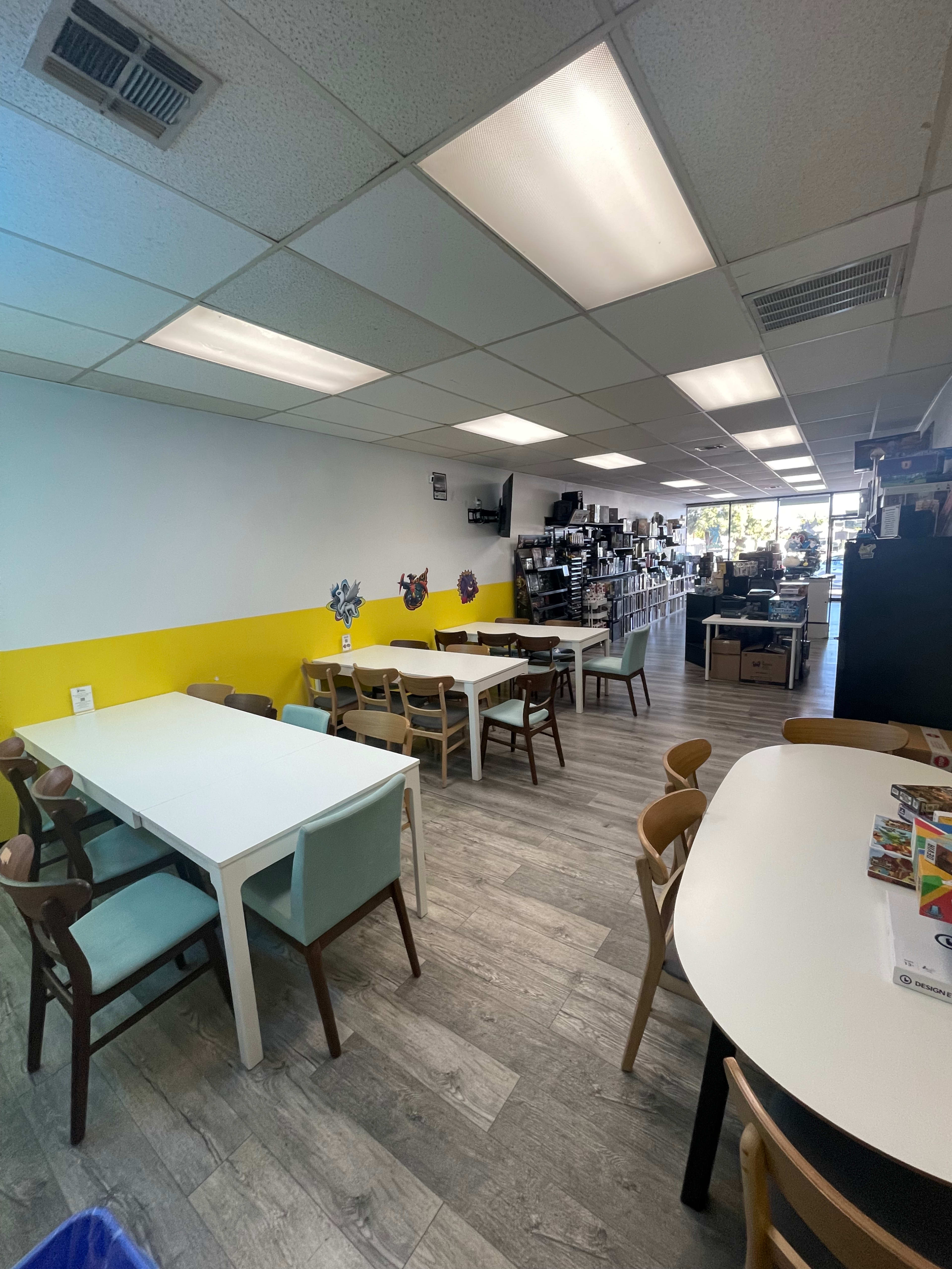 A spacious room with multiple tables and chairs, featuring a yellow accent wall and shelves filled with books and materials.