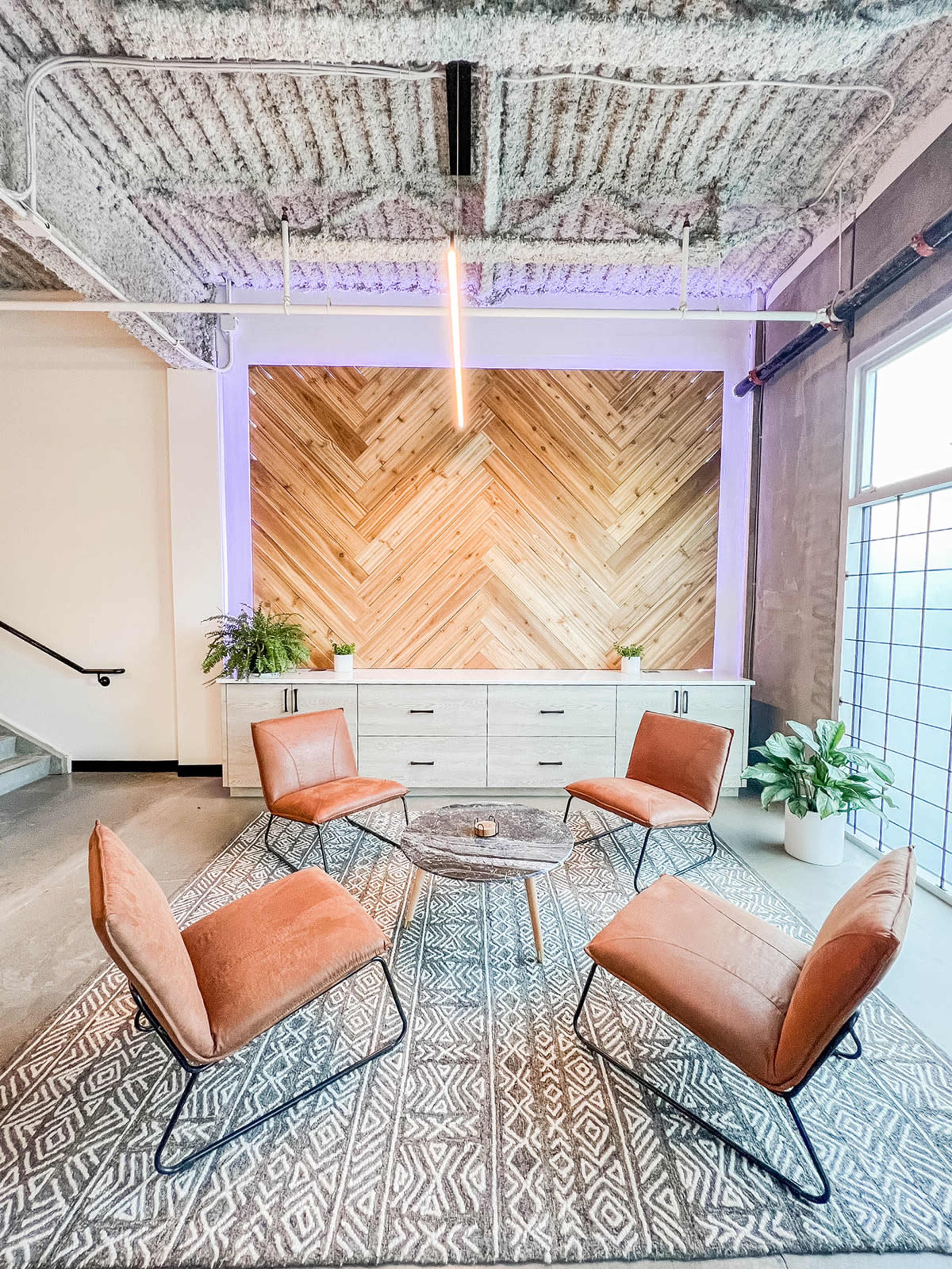 A modern lounge area features four brown chairs arranged around a circular coffee table on a patterned rug in front of a wooden accent wall.