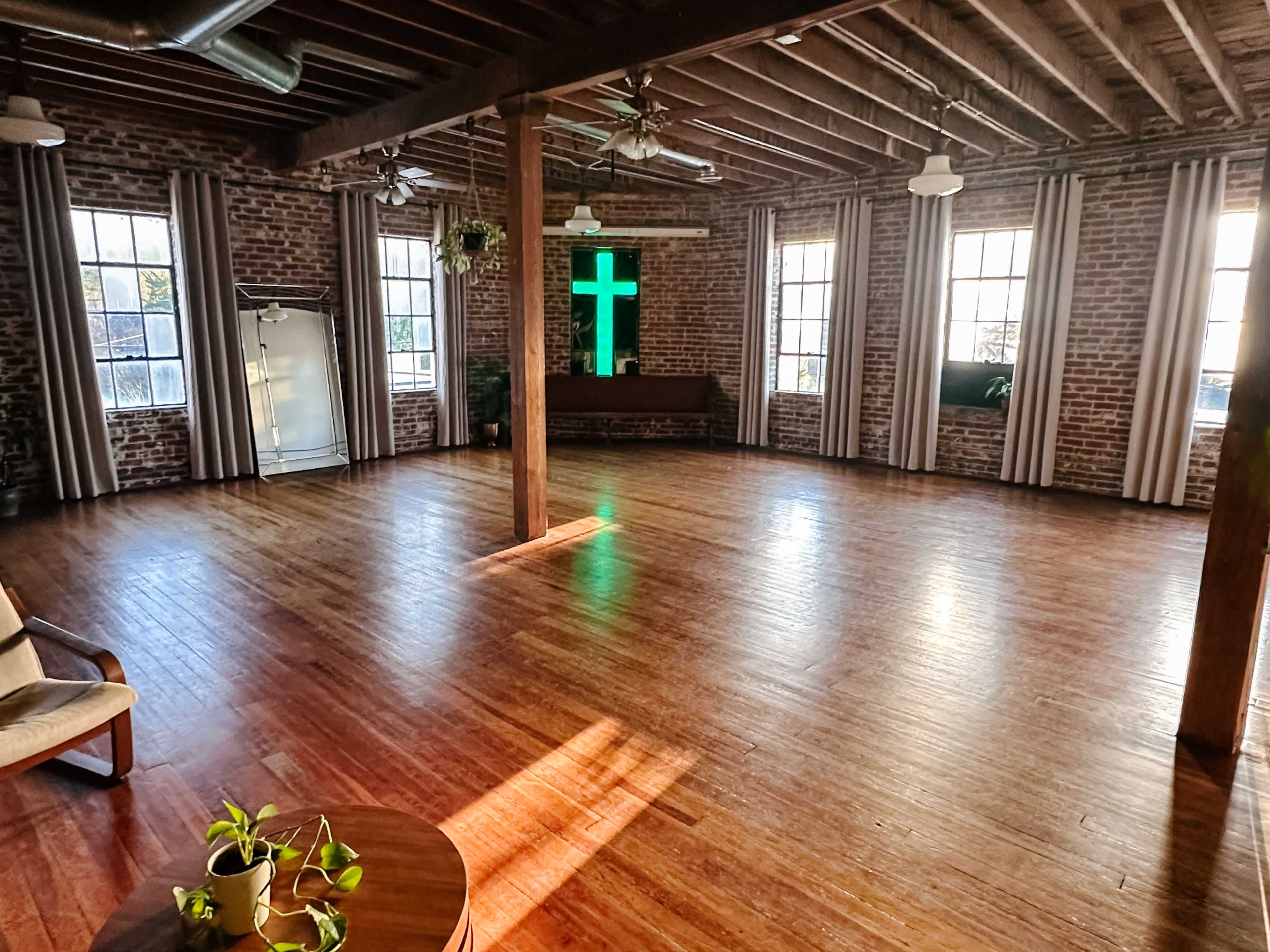 The image shows a spacious room with wooden floors, exposed brick walls, and large windows, featuring a green cross illuminated by natural light.