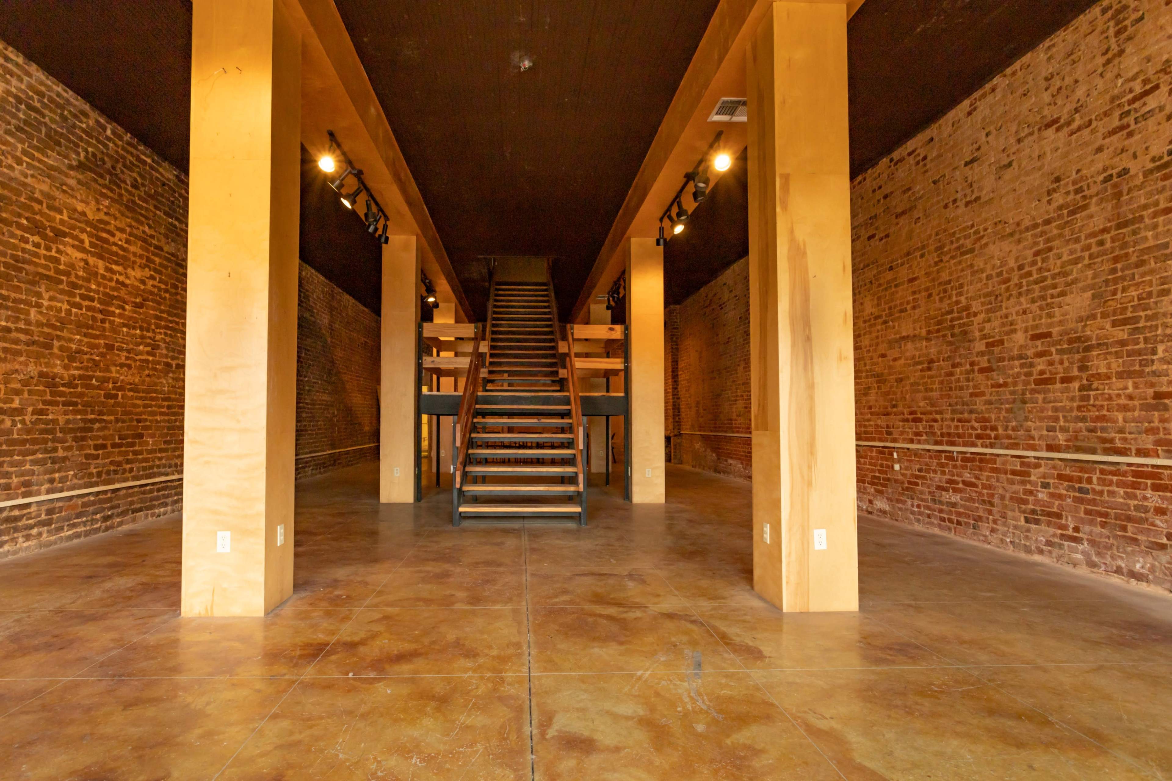 The image shows an empty interior space featuring exposed brick walls, polished concrete flooring, and a staircase at the back.