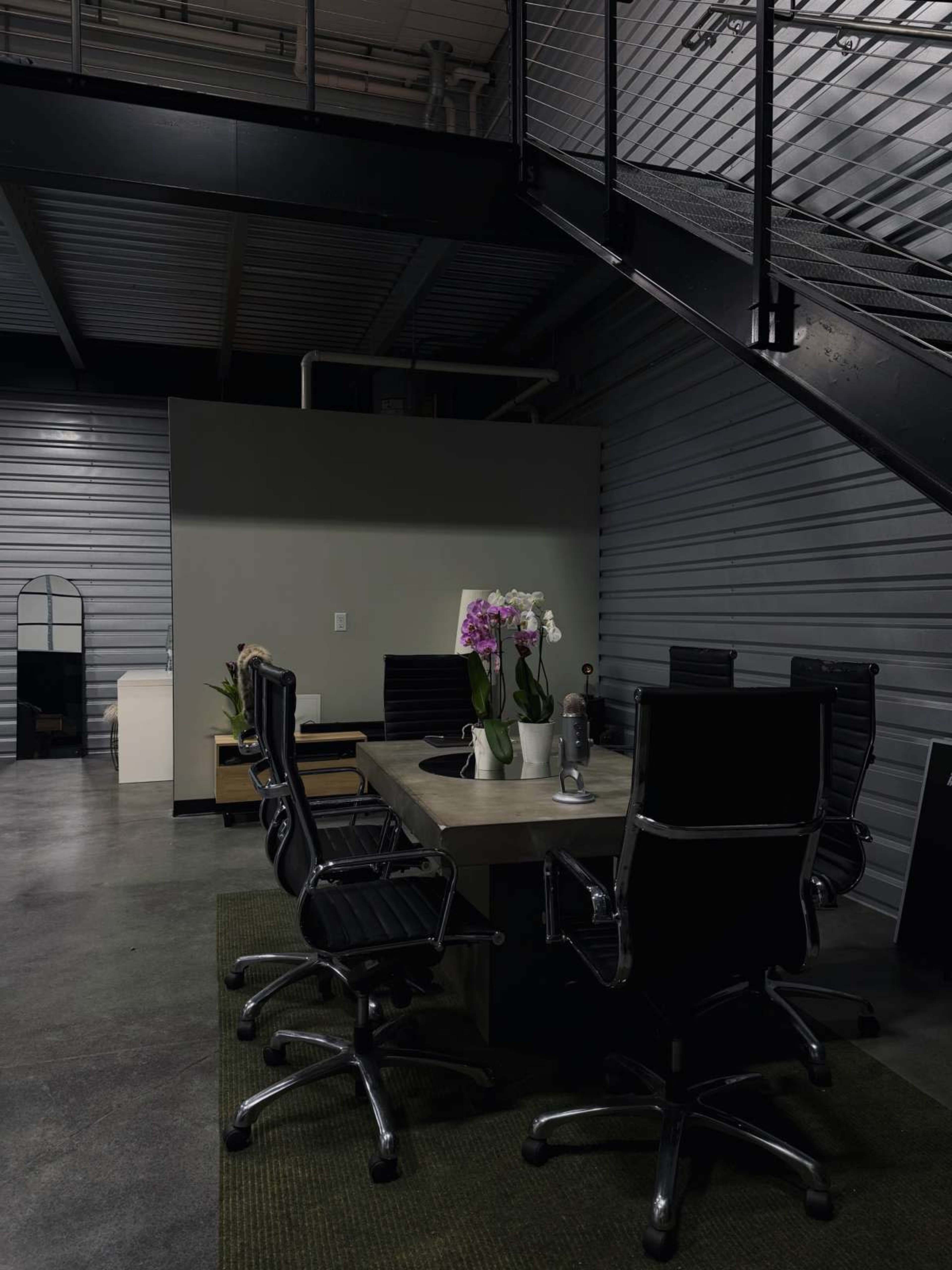 A conference room with a rectangular table surrounded by black chairs, featuring a vase of orchids in the center, and a staircase visible in the background.