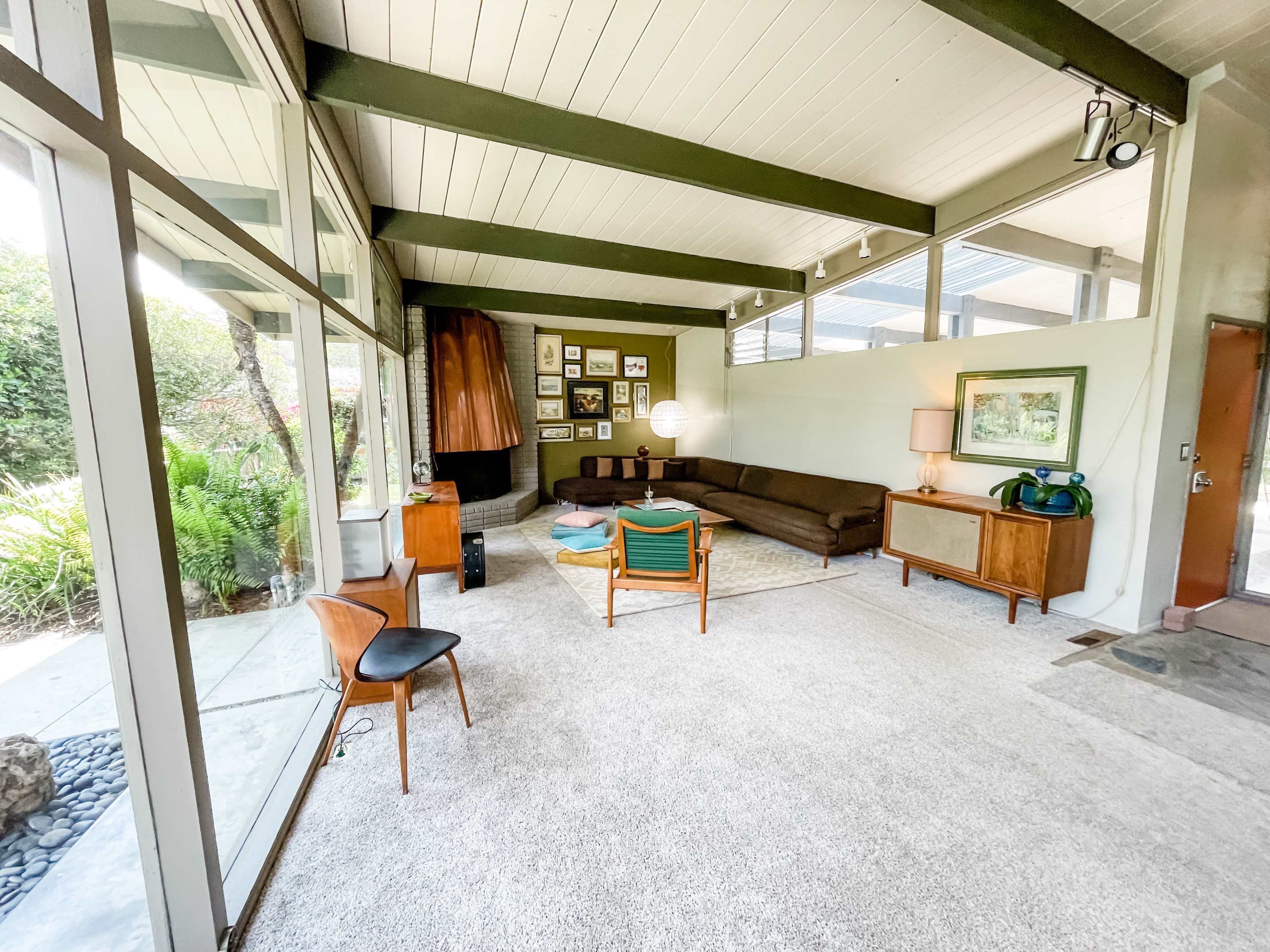The image shows a modern living room with large glass windows, a brown sectional sofa, a wooden side table, and a gallery wall of framed artwork.