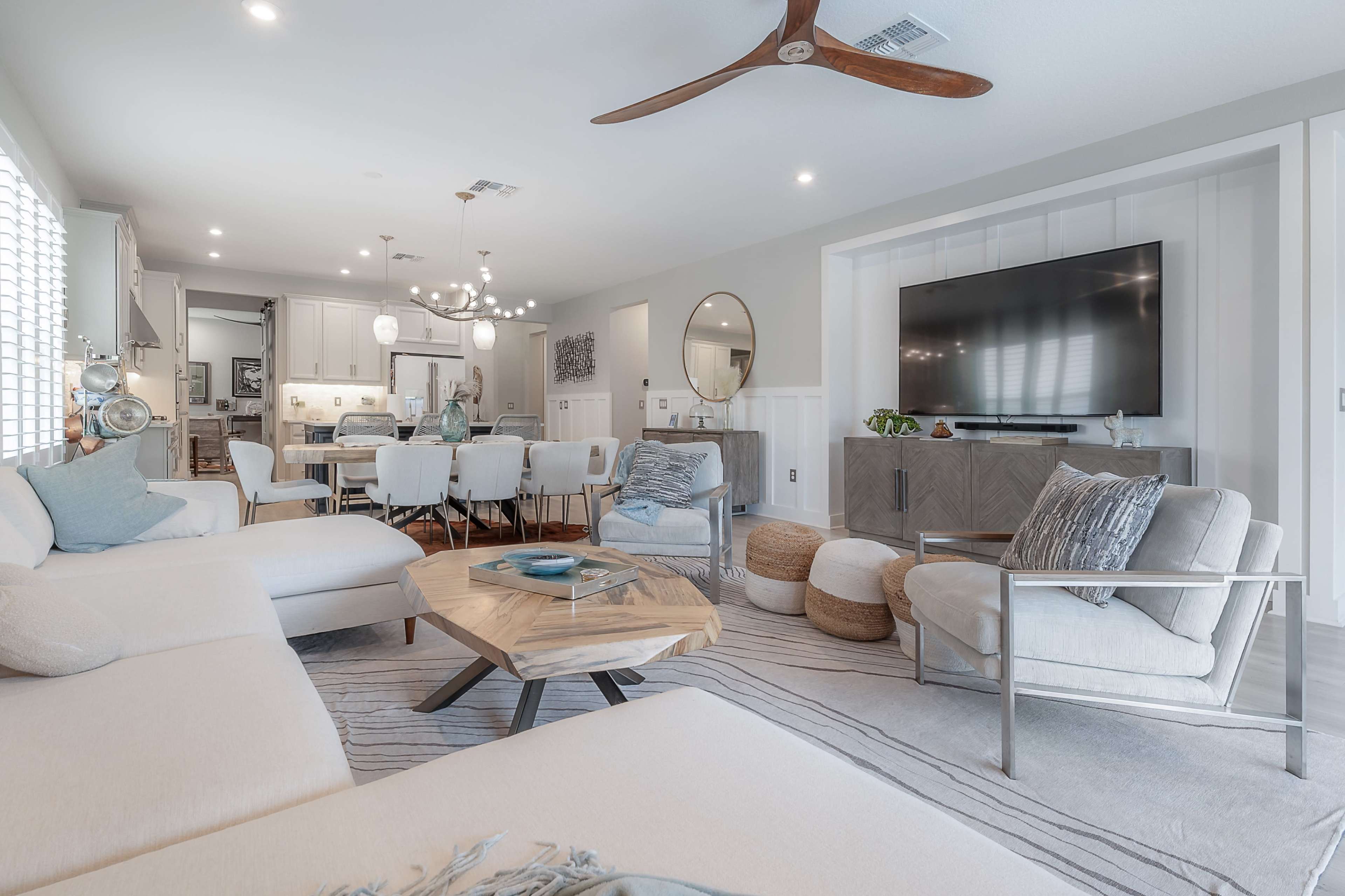 The living room features a light-colored sectional sofa, a wooden coffee table, and a large television mounted on the wall, with a dining area visible in the background.