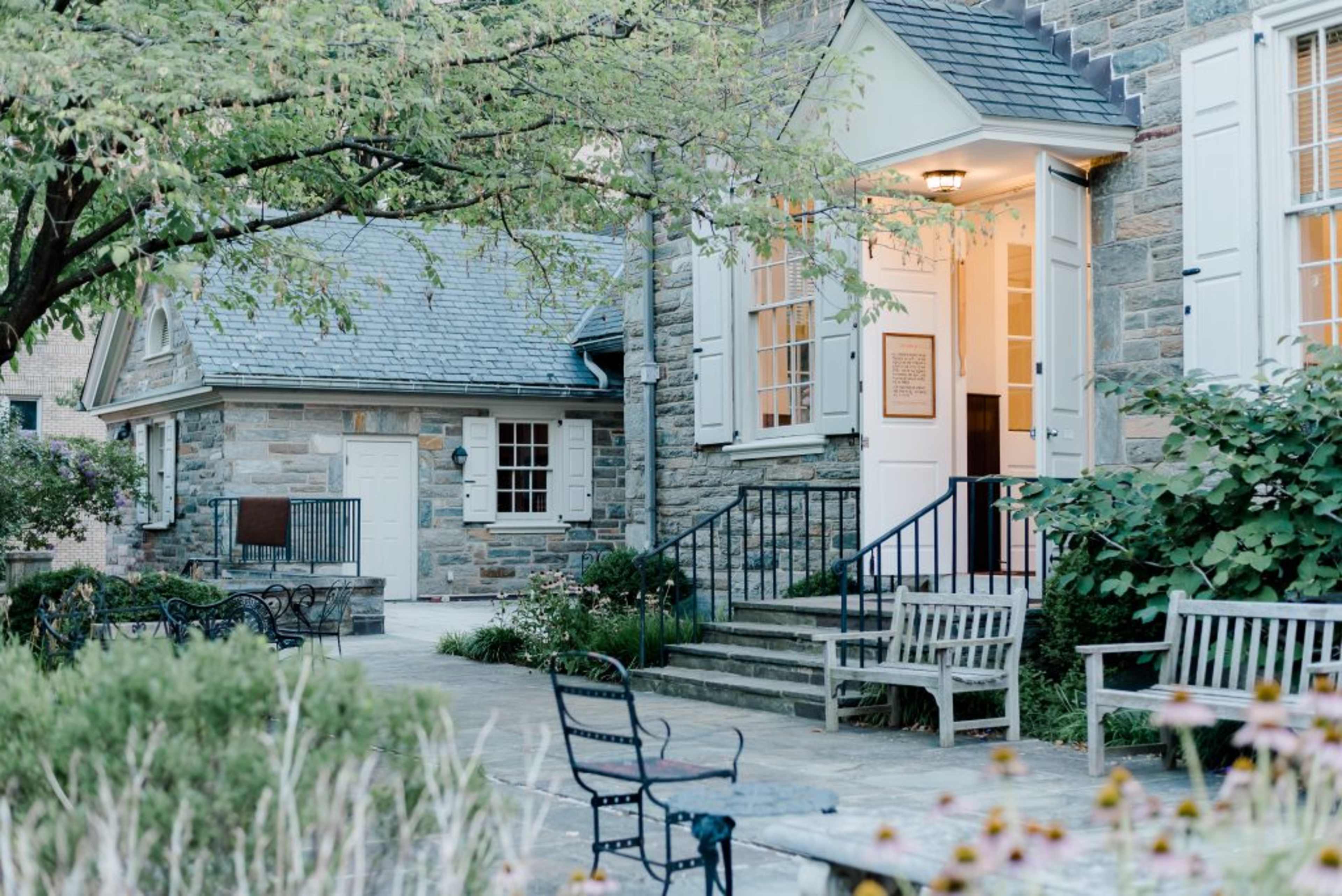 The image shows a stone building with white shutters and an entrance flanked by benches and greenery.