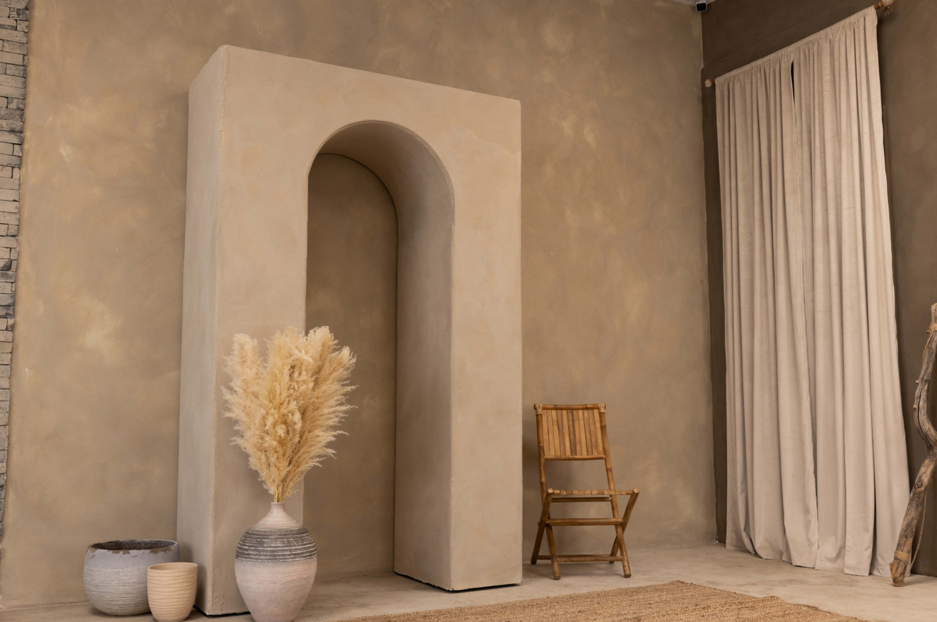 A minimalist interior space featuring a large archway, a vase with dried grasses, a wooden chair, and neutral-colored walls and curtains.