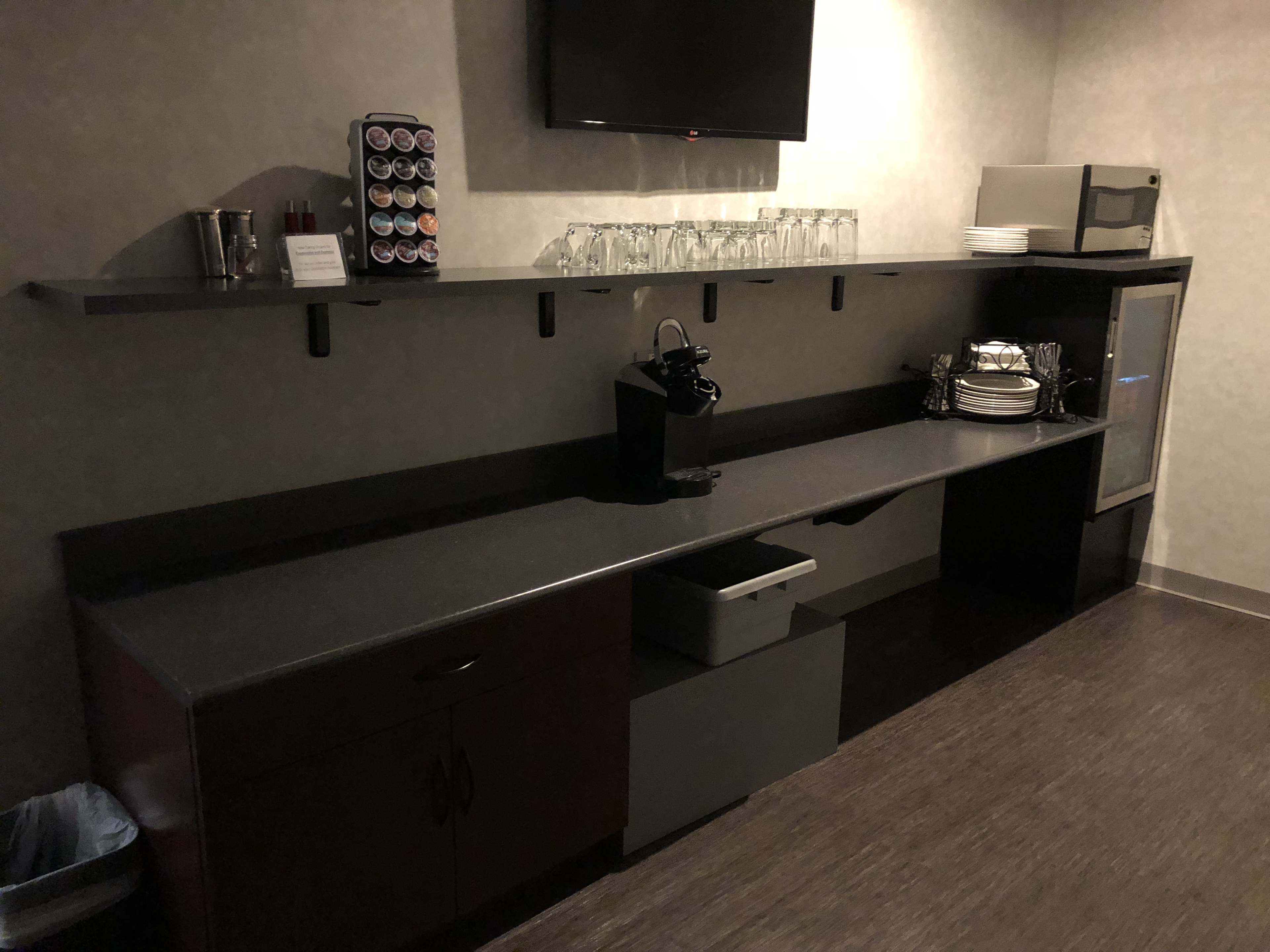 The image shows a modern countertop area featuring a coffee maker, a shelf with glasses and coffee pods, and a small refrigerator beneath.