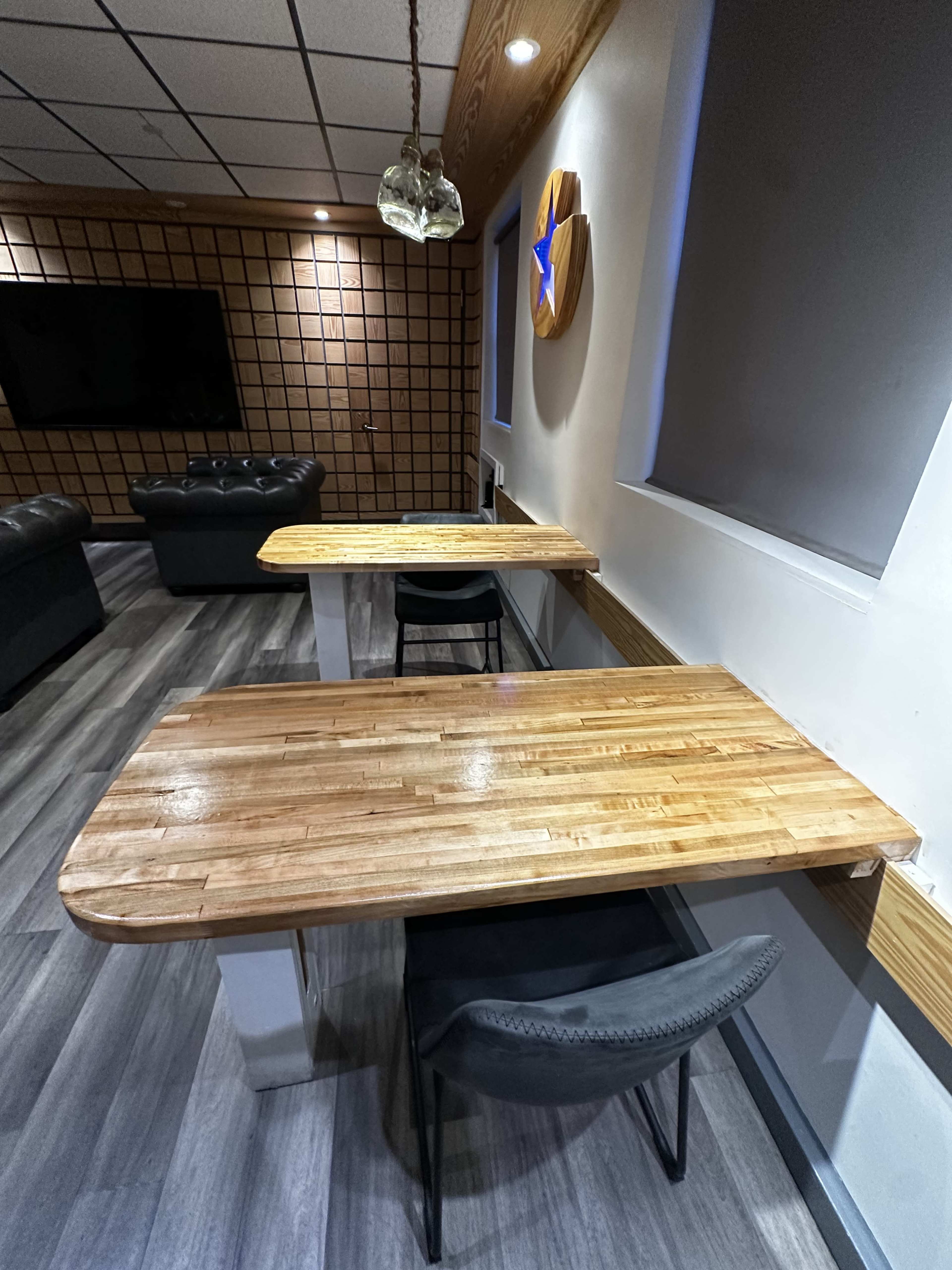 The image shows two wooden tables with chairs in a room featuring a textured wall and a television on the opposite side.