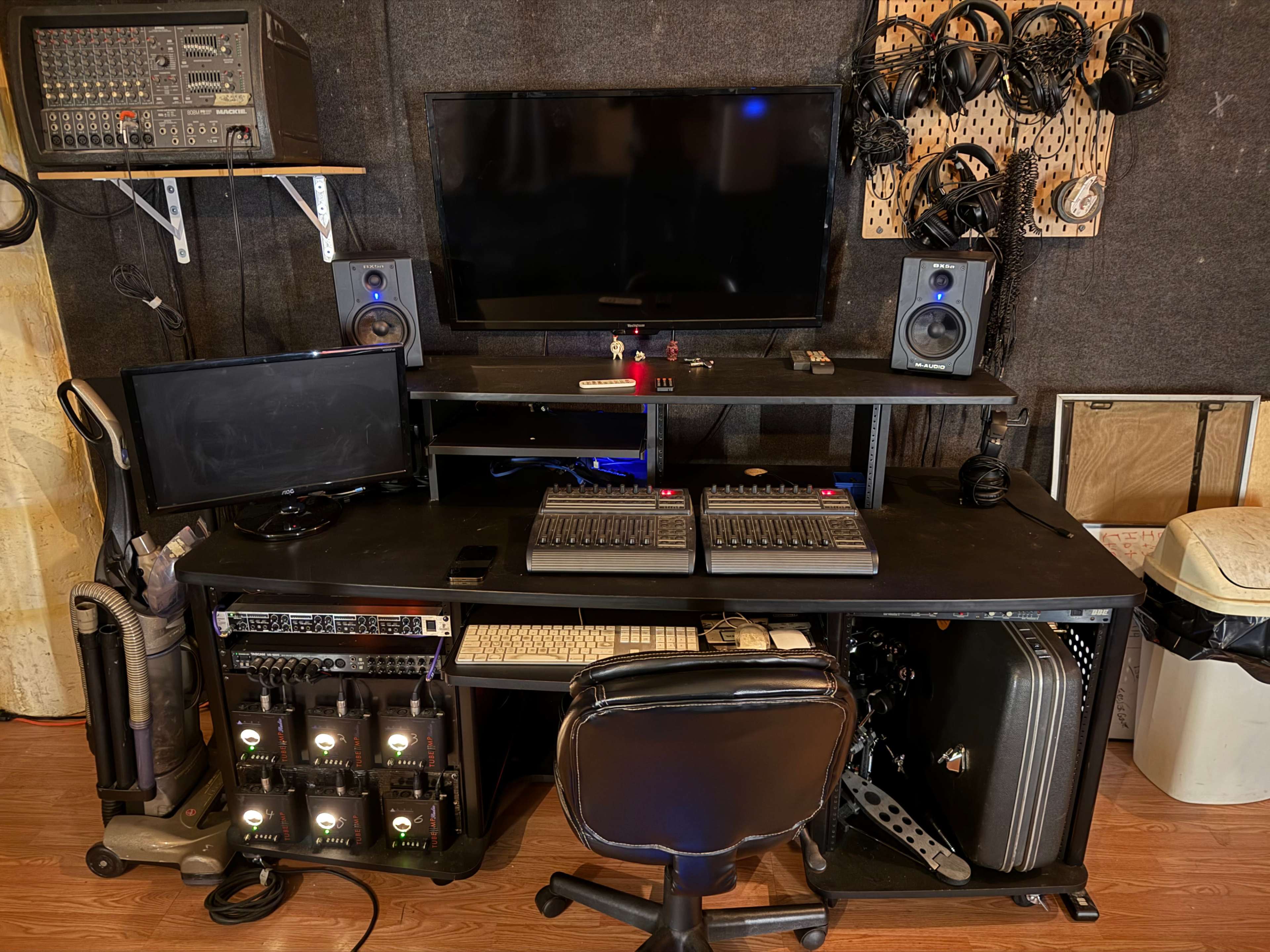 The image shows a music production setup with a large desk containing audio equipment, two monitors, a mixer, speakers, and organized headphones hanging on the wall.