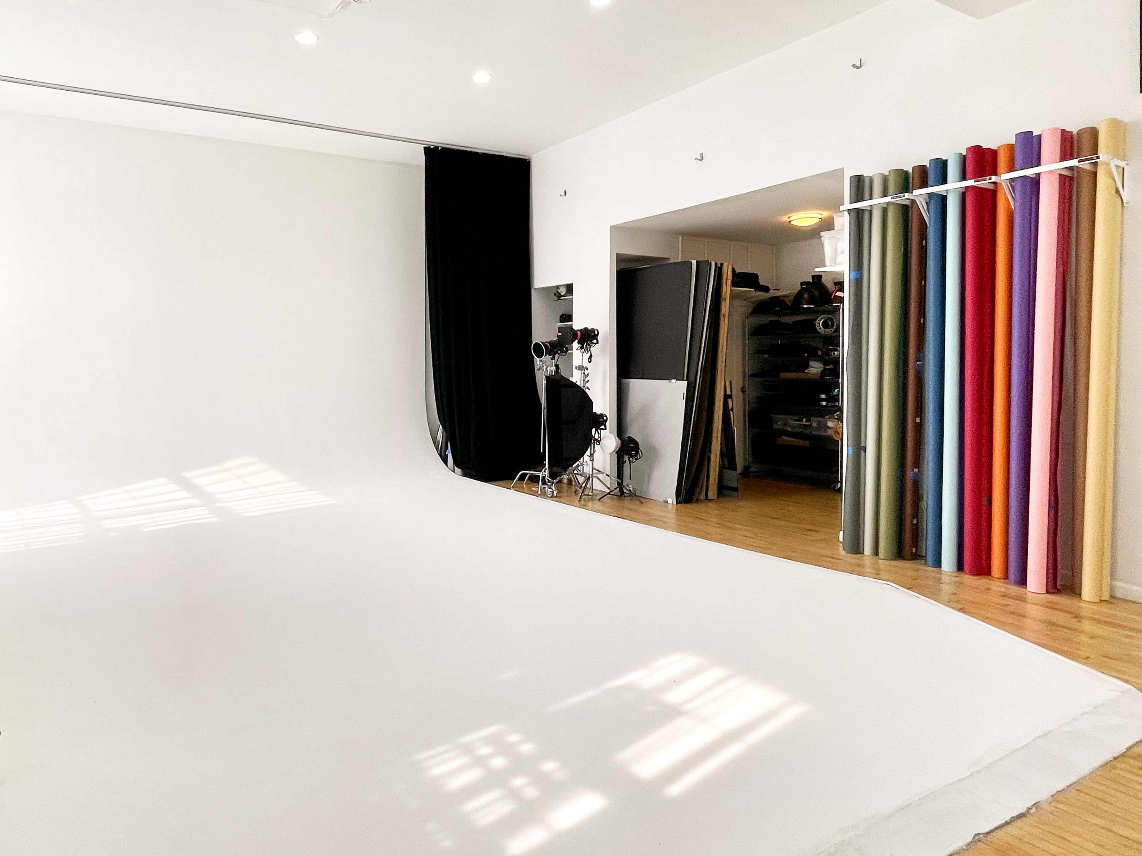 A spacious photography studio with a white seamless backdrop, studio lights on the left, and a storage area featuring rolls of colored backdrops on the right.