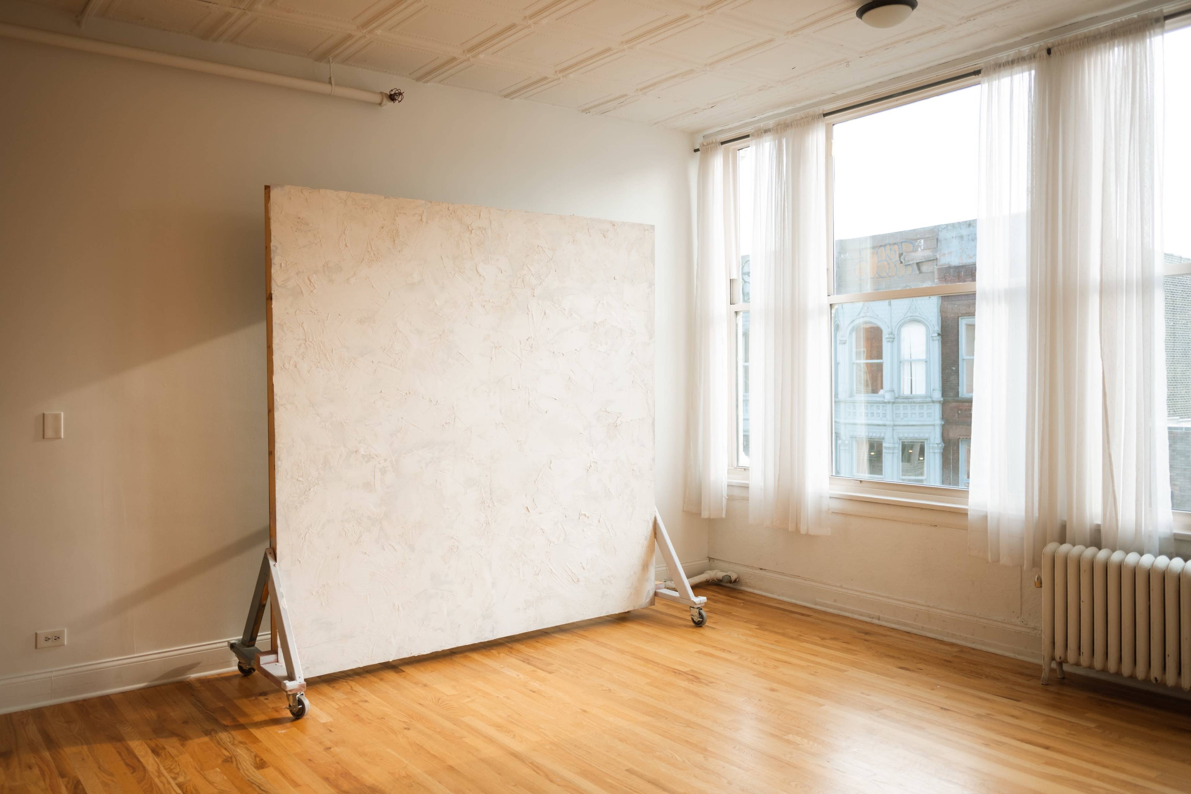 A spacious, well-lit room featuring a large, unpainted canvas on wheels positioned against a wall with large windows.