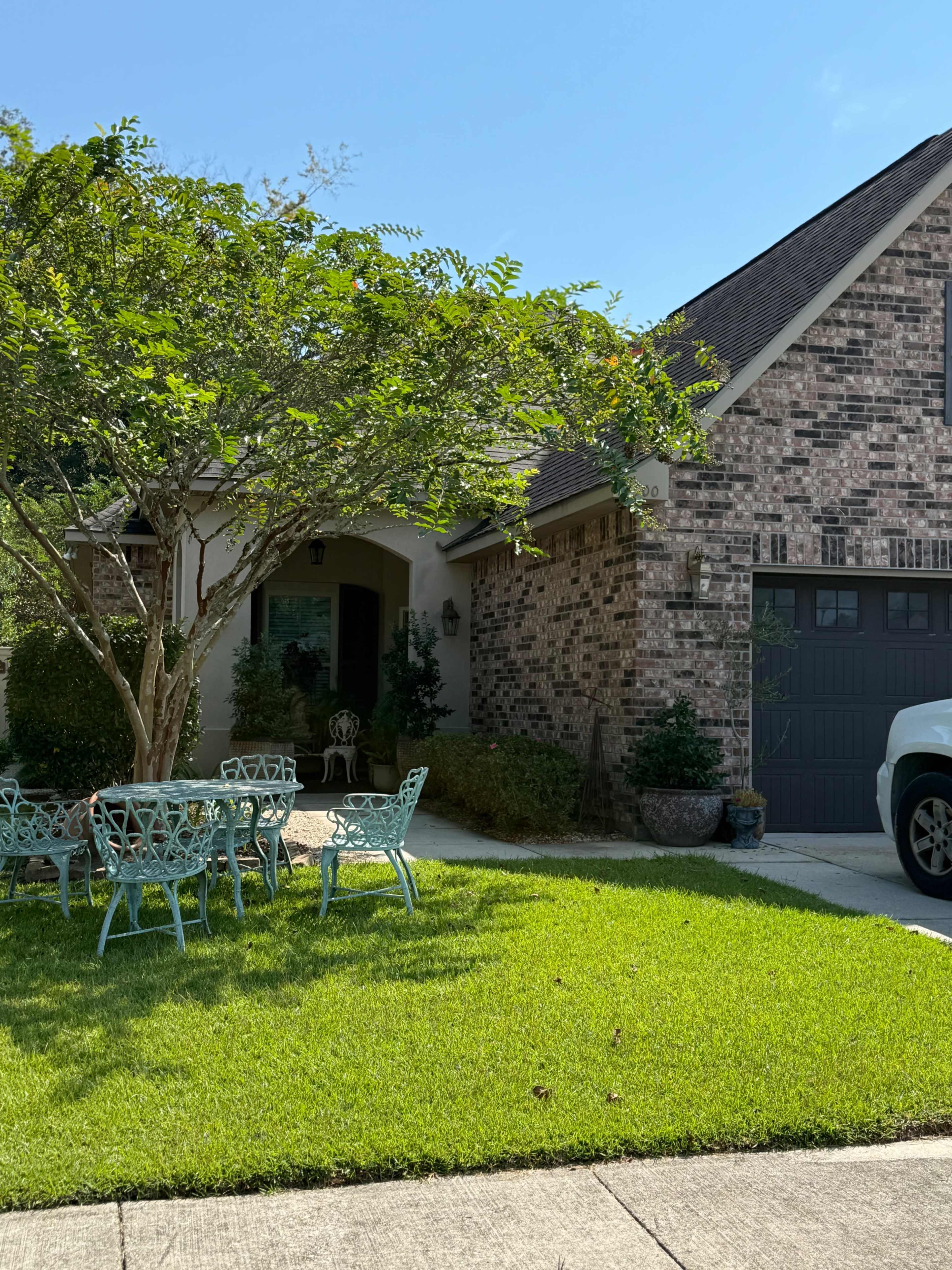The image shows a brick house with a manicured lawn, a small patio set, and a vehicle parked in the driveway.