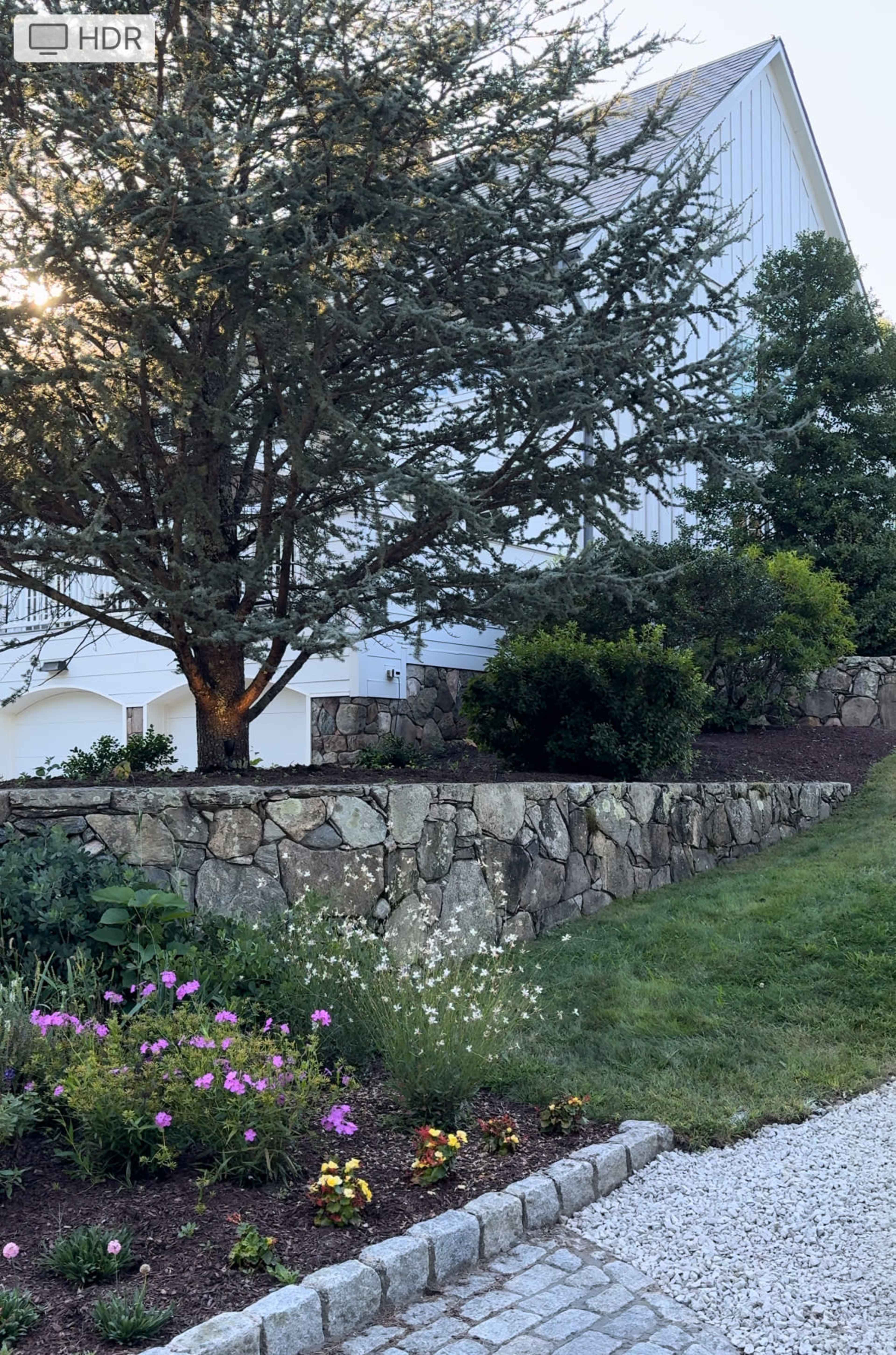 A stone retaining wall supports a landscaped garden at the base of a house, featuring a tree and various flowering plants.