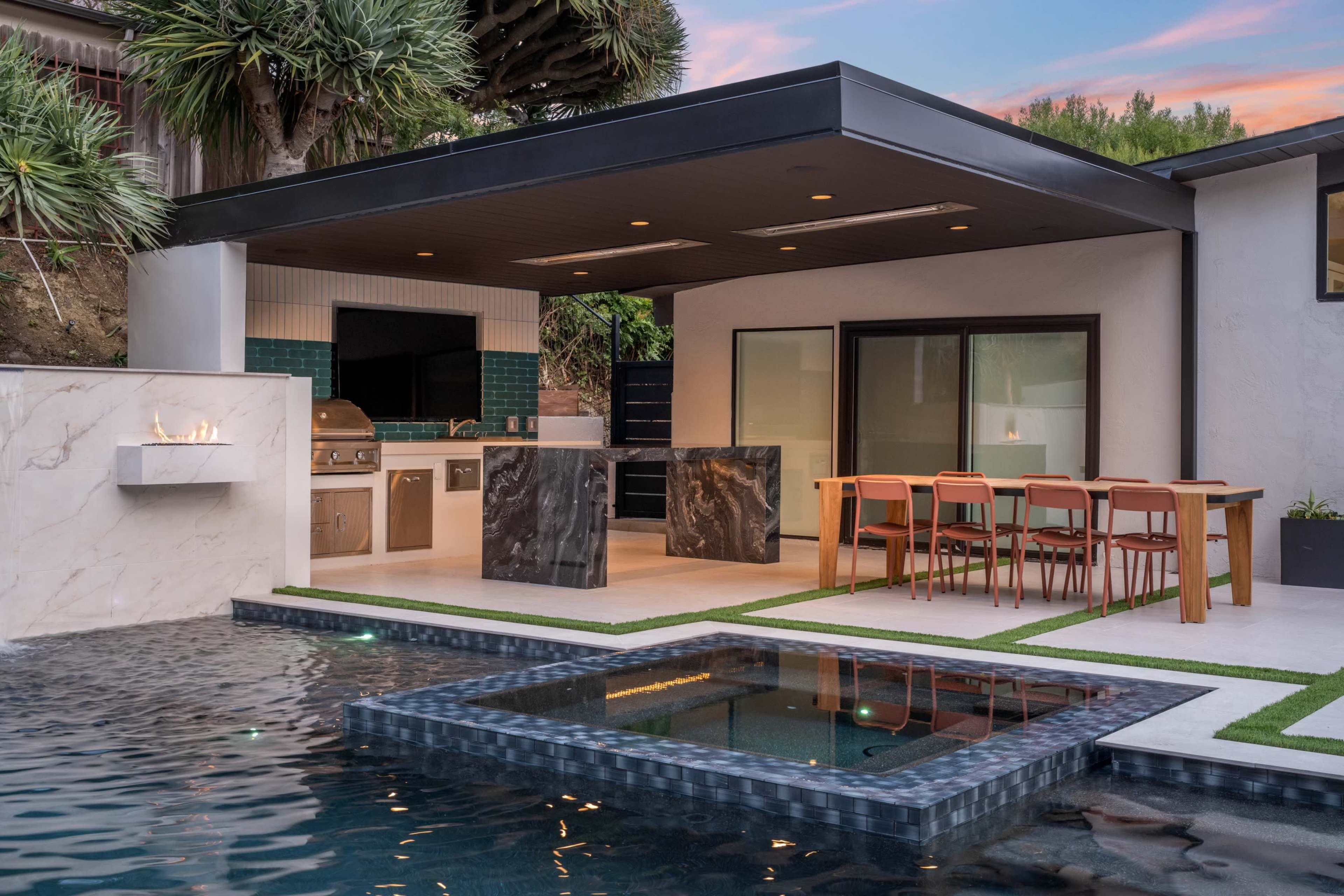The image shows a modern outdoor kitchen with a marble island and seating area, adjacent to a swimming pool with a sunken hot tub surrounded by greenery.