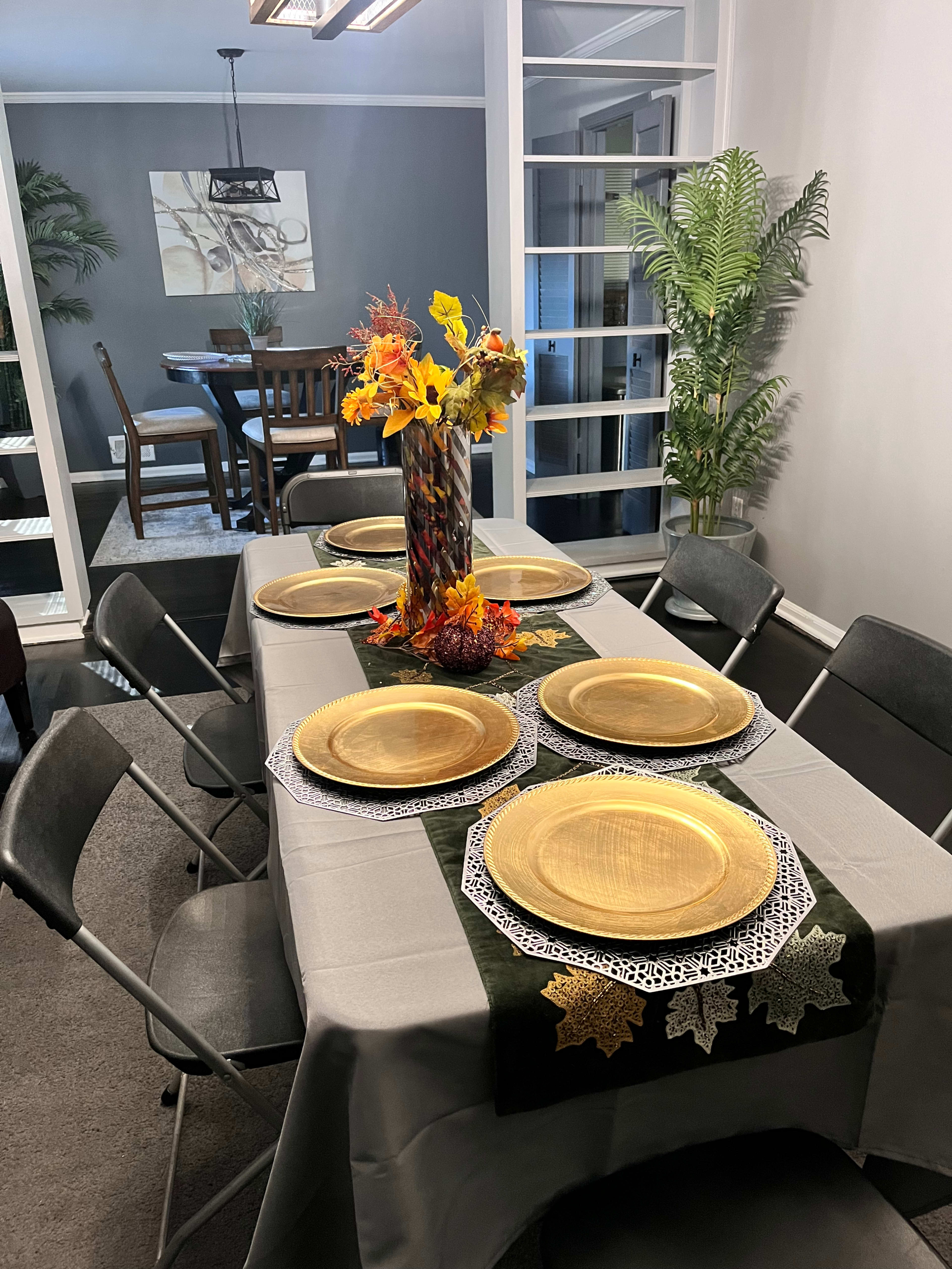 A formal dining table is set with gold plates and a centerpiece of autumn flowers, while covered by a gray tablecloth and surrounded by folding chairs.
