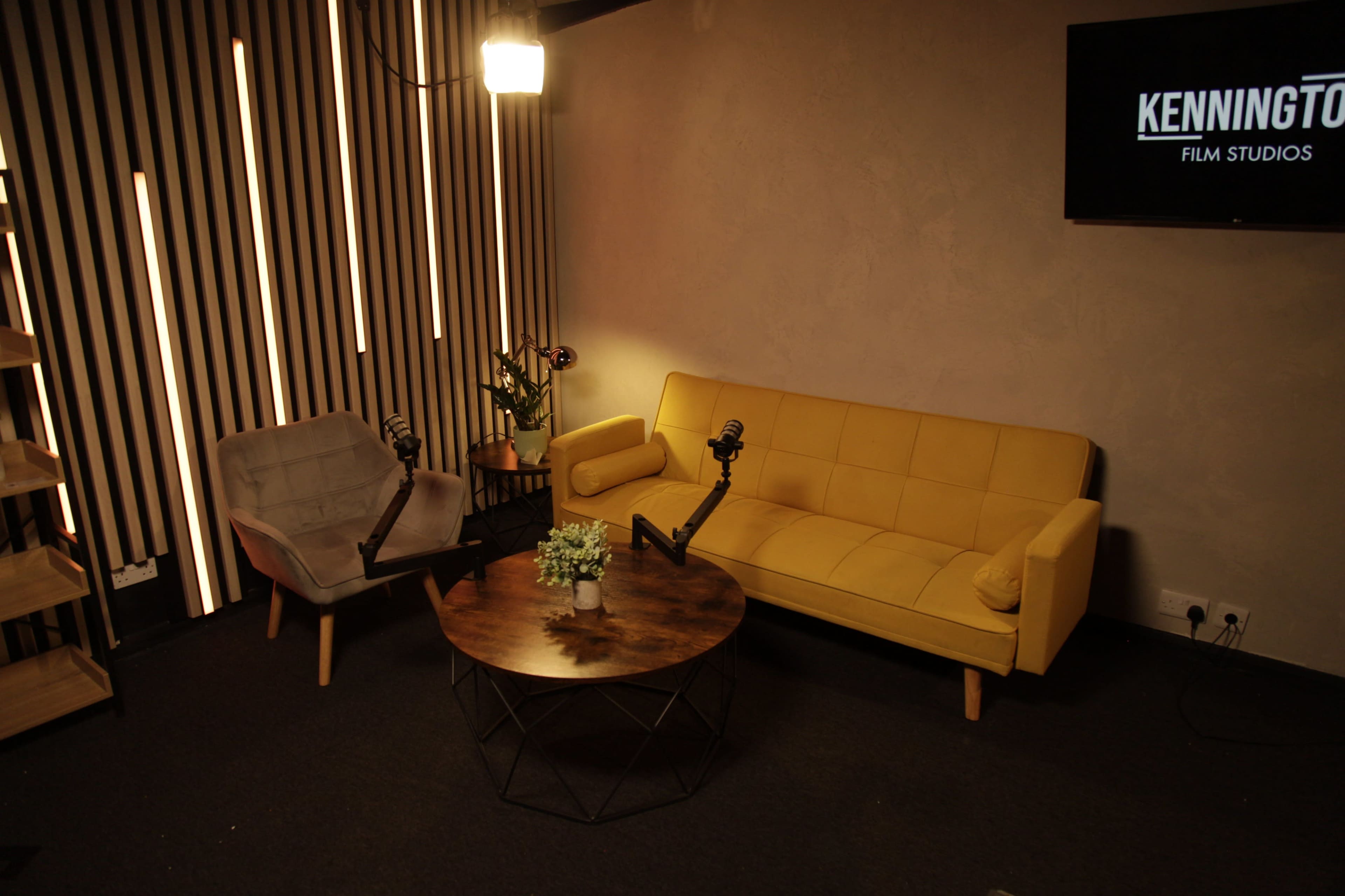 A modern studio setting features a yellow sofa, gray armchairs, and a circular table, with microphones positioned for recording.