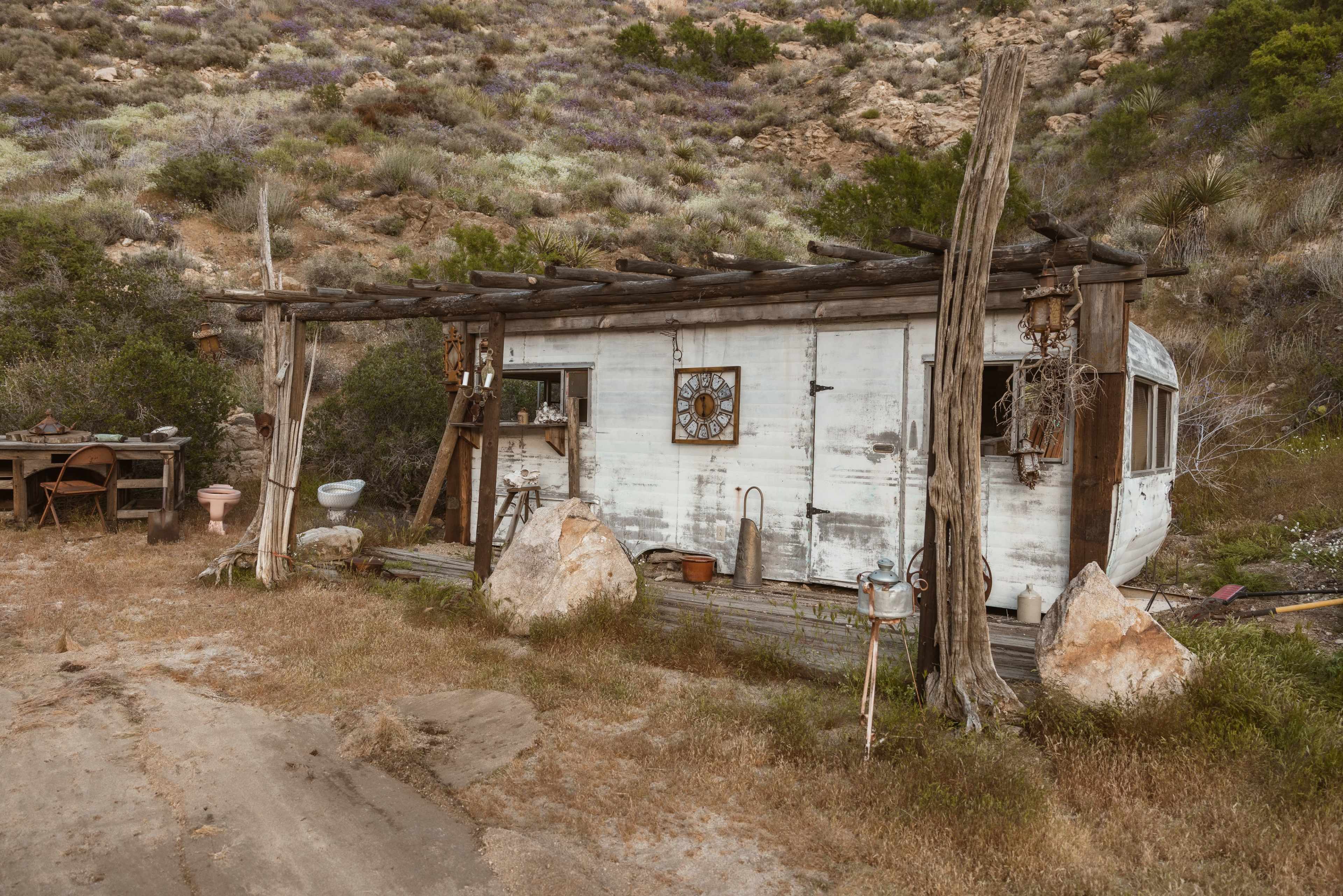 A weathered, small trailer house with a wooden overhang is situated among rocky terrain and sparse vegetation.
