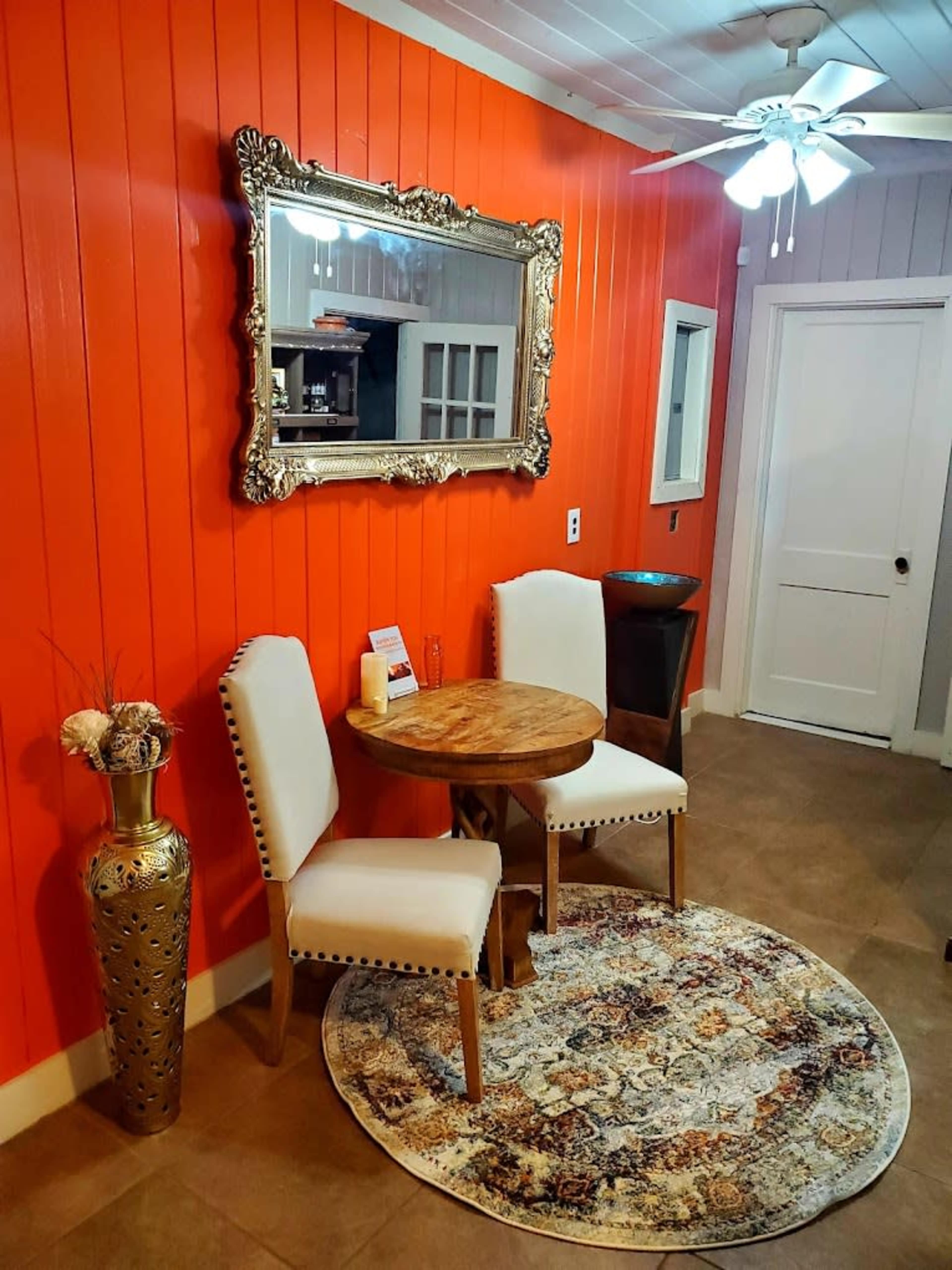 A small dining area features a round wooden table with two upholstered chairs, set against an orange wall and accompanied by a decorative mirror and a rug.