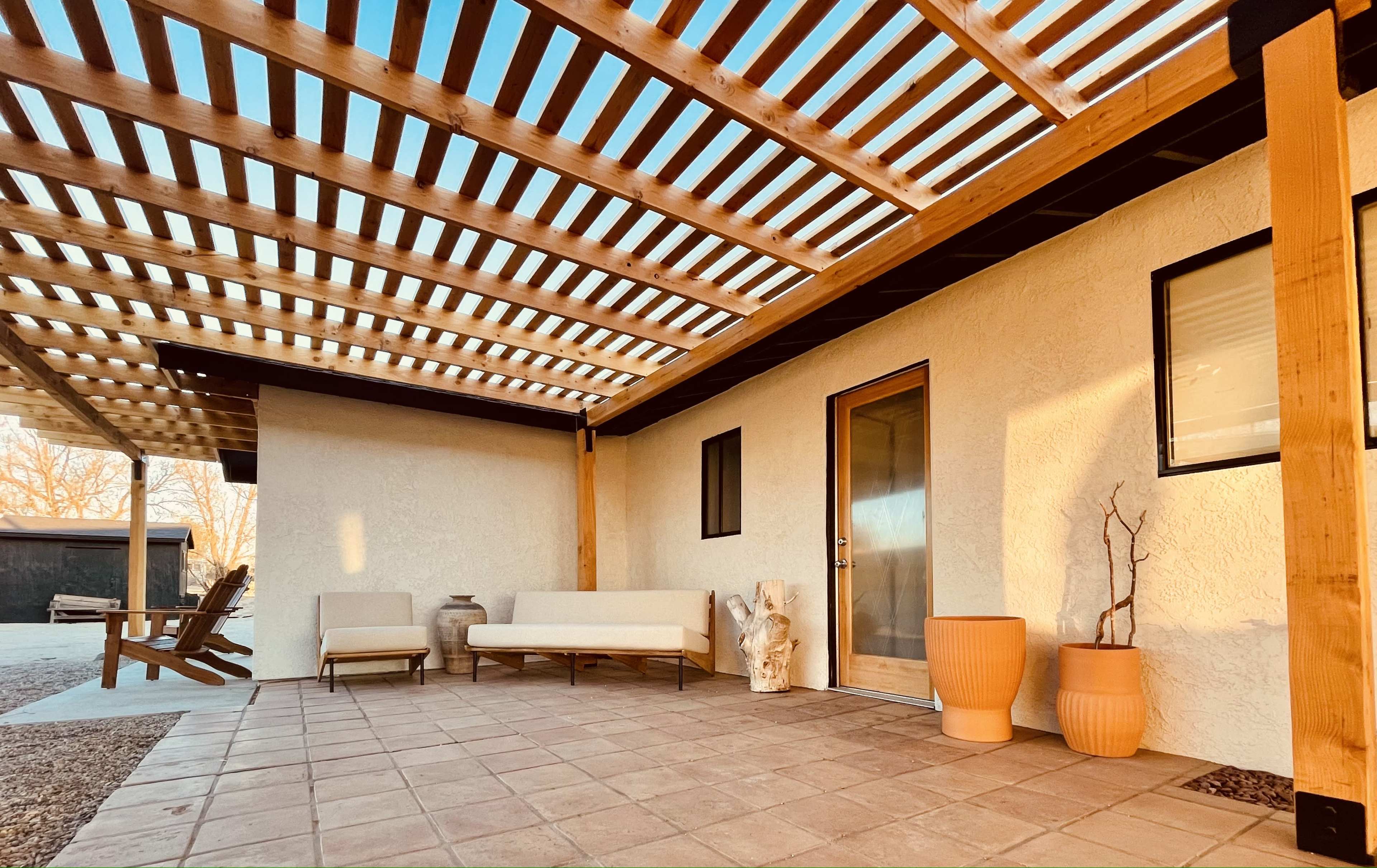 A patio area sheltered by a wooden pergola, featuring a sofa, decorative planters, and a door leading into a house.