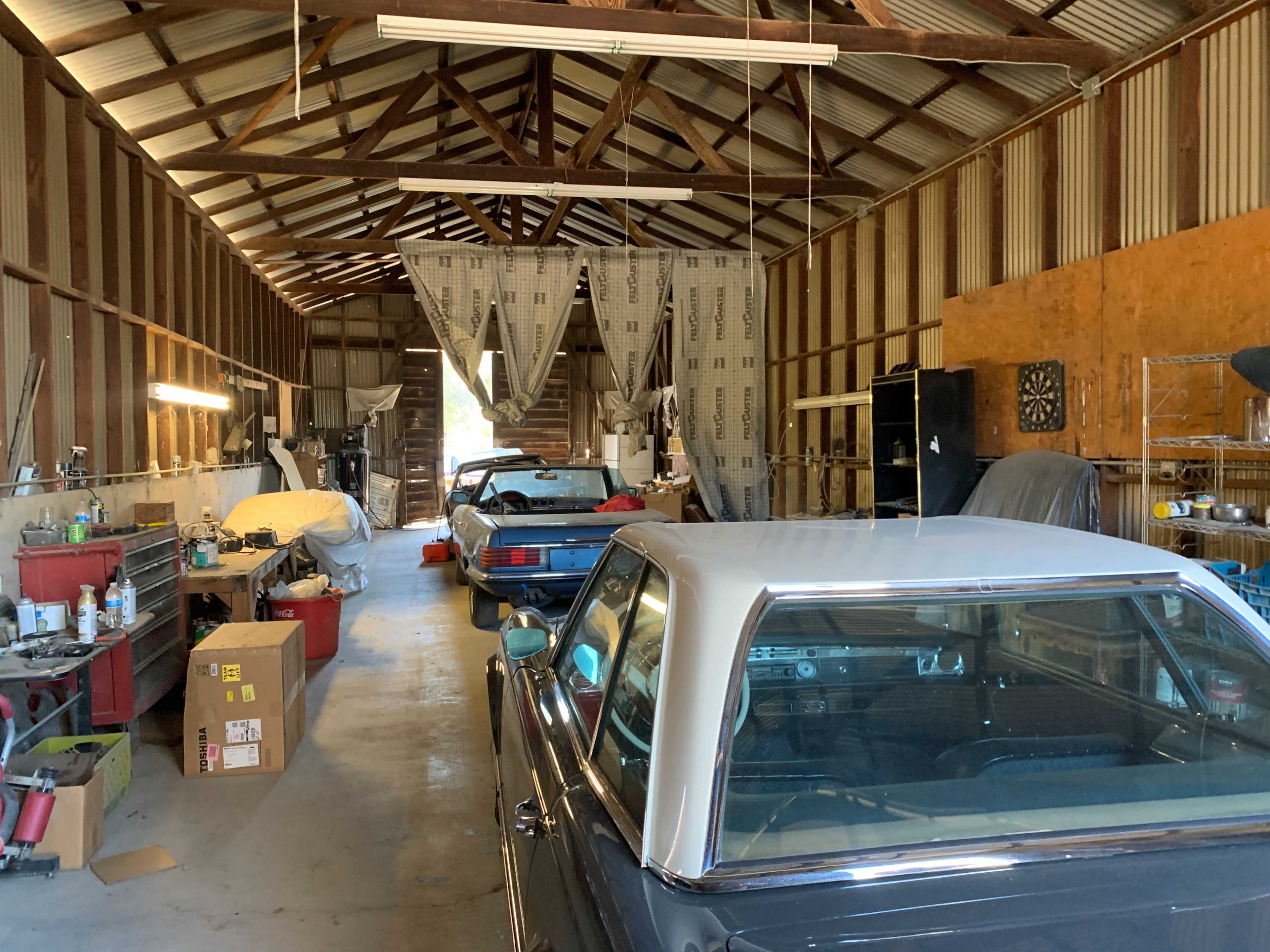 The image shows a spacious workshop with classic cars, tools, and supplies organized along the walls in a storage building.